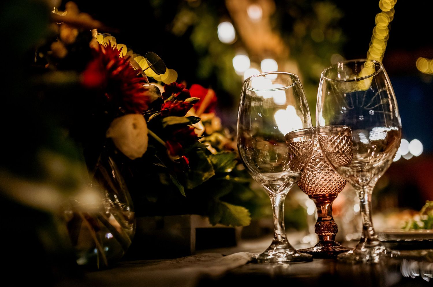 Elegant crystal wine glasses on a table with bokeh lights and red floral arrangement at a romantic dinner setting.