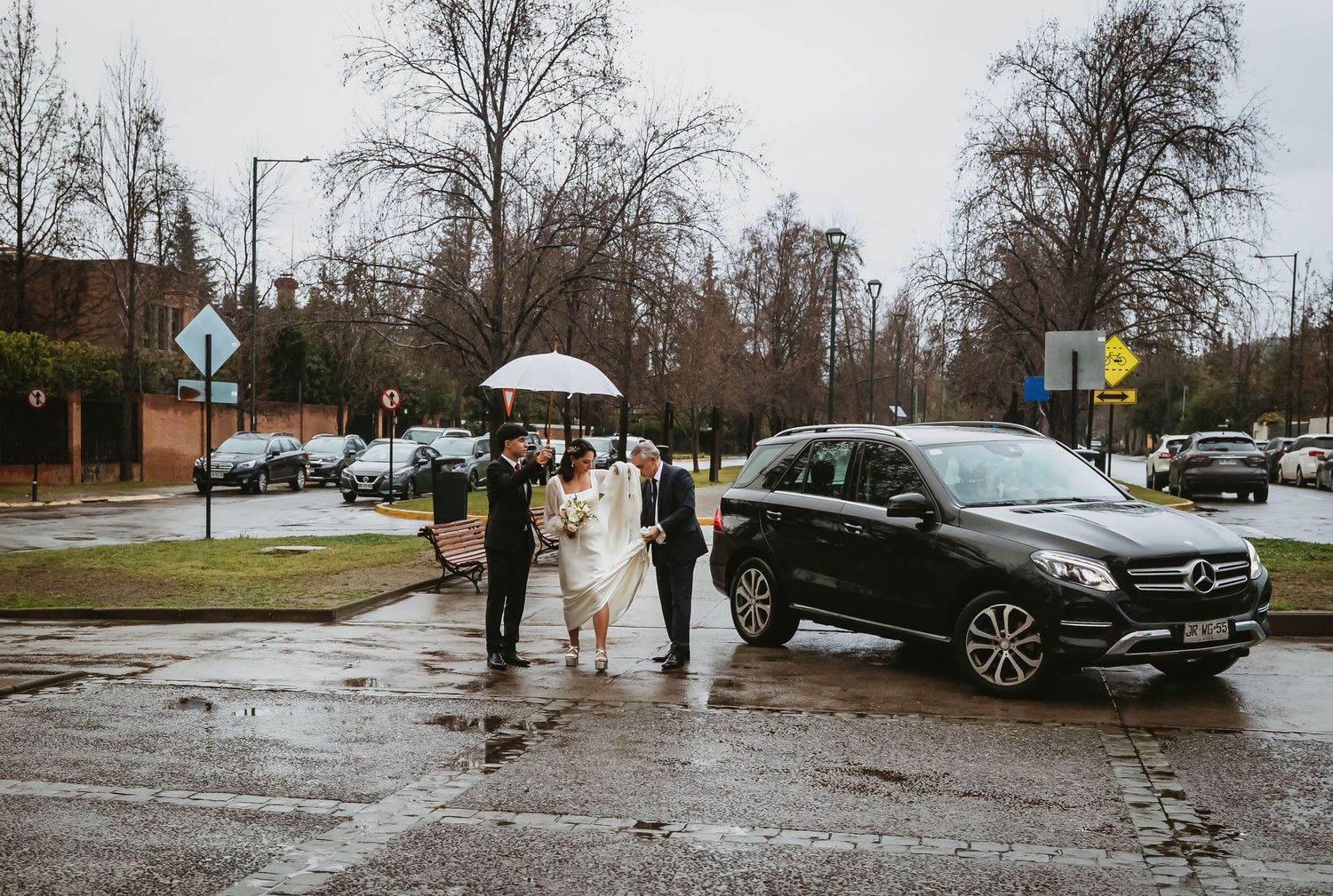 Wedding couple with guests under umbrella on rainy day beside a Mercedes SUV on a wet street.