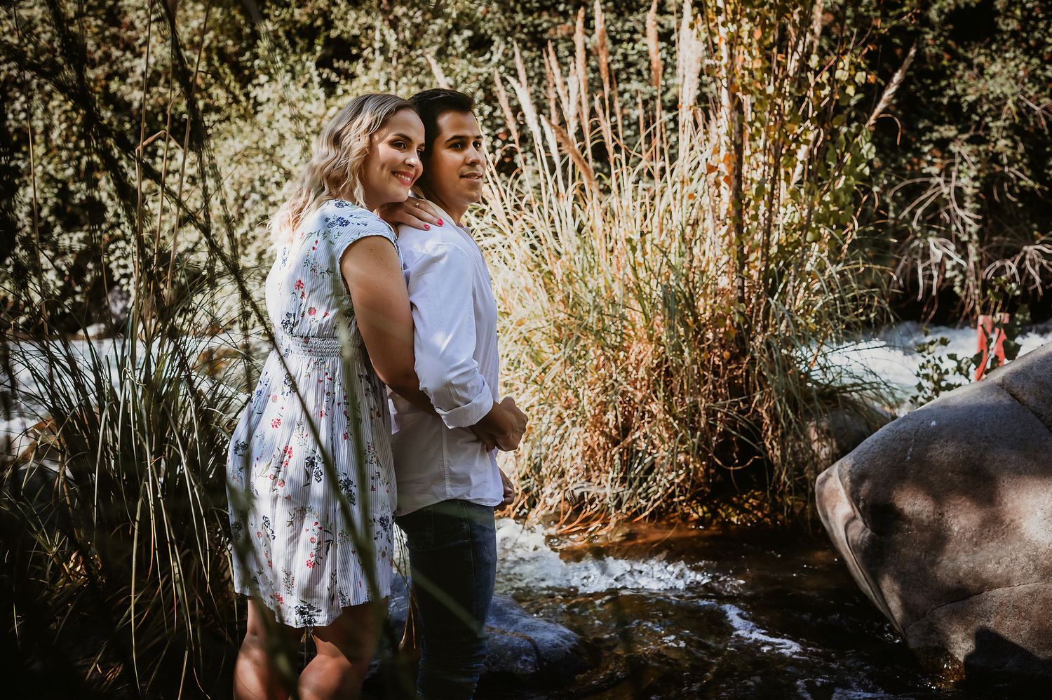 Couple embracing outdoors near a stream surrounded by lush greenery and tall grasses in a romantic nature setting.