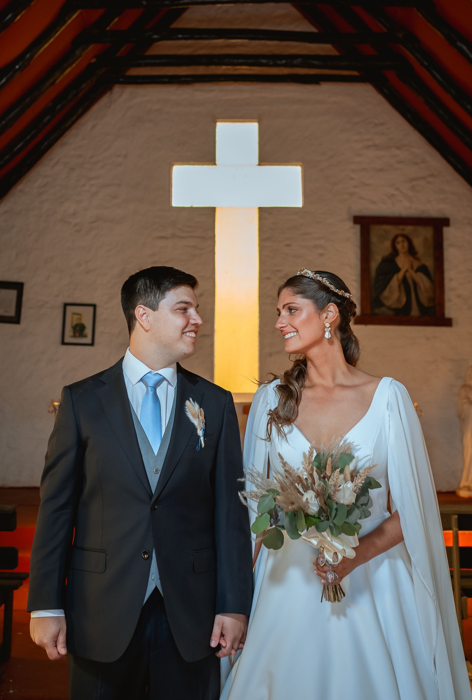 Bride and groom exchange loving glances inside a rustic chapel with an illuminated cross backdrop on their wedding day.