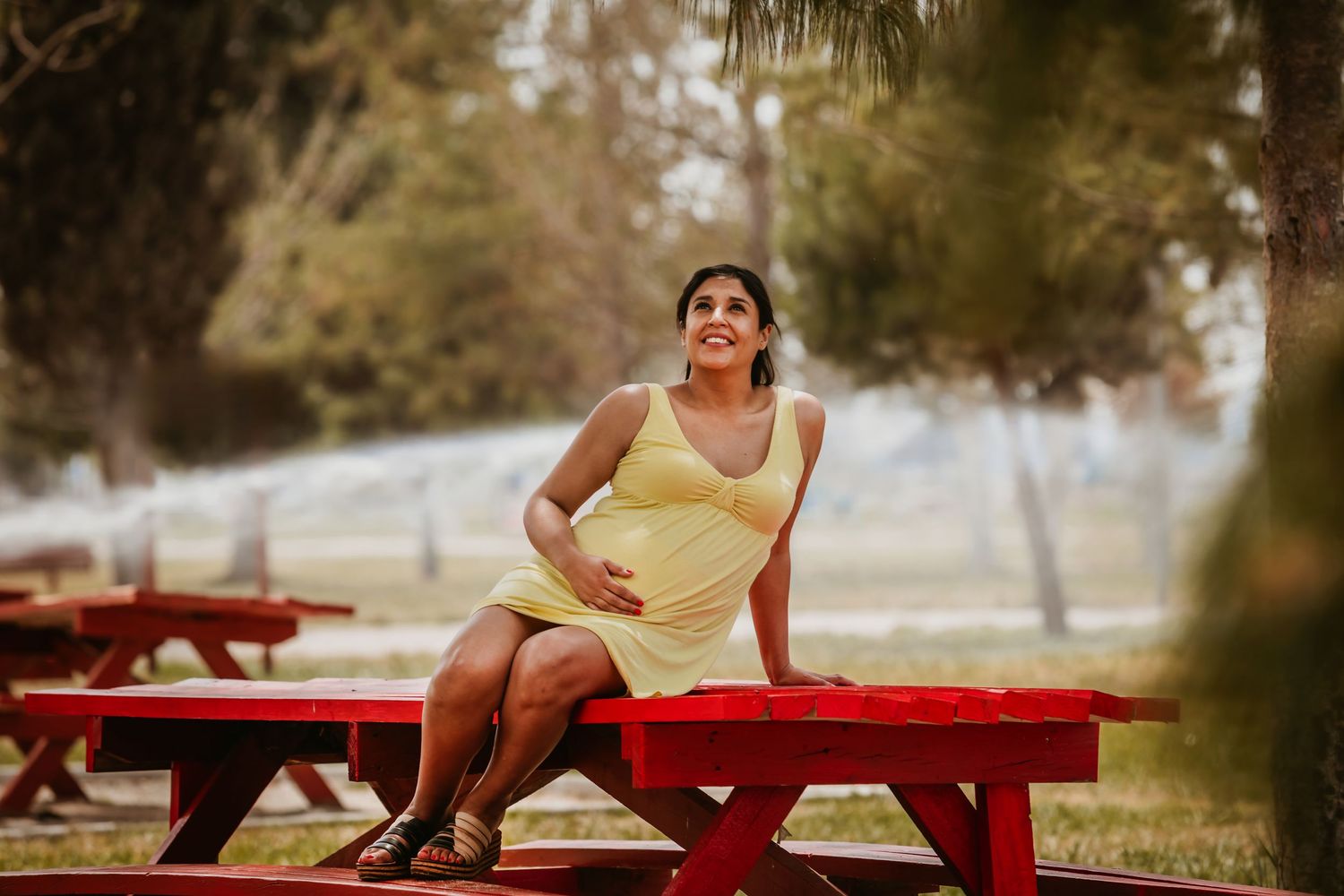 Pregnant woman in yellow dress sitting on red picnic table, smiling outdoors in a park setting.