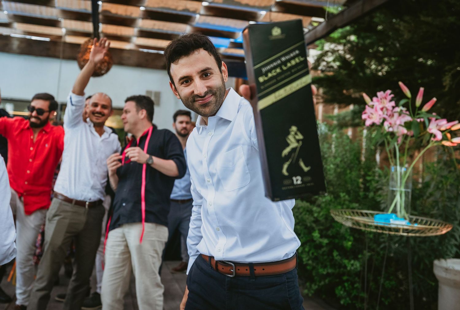 Man smiling holding Johnnie Walker Black Label whisky bottle at outdoor celebration party with guests in background.