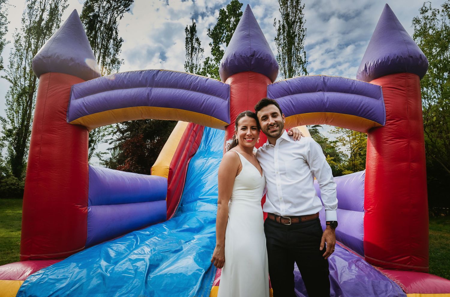 Smiling bride and groom posing in front of a colorful inflatable bounce castle at an outdoor wedding reception.