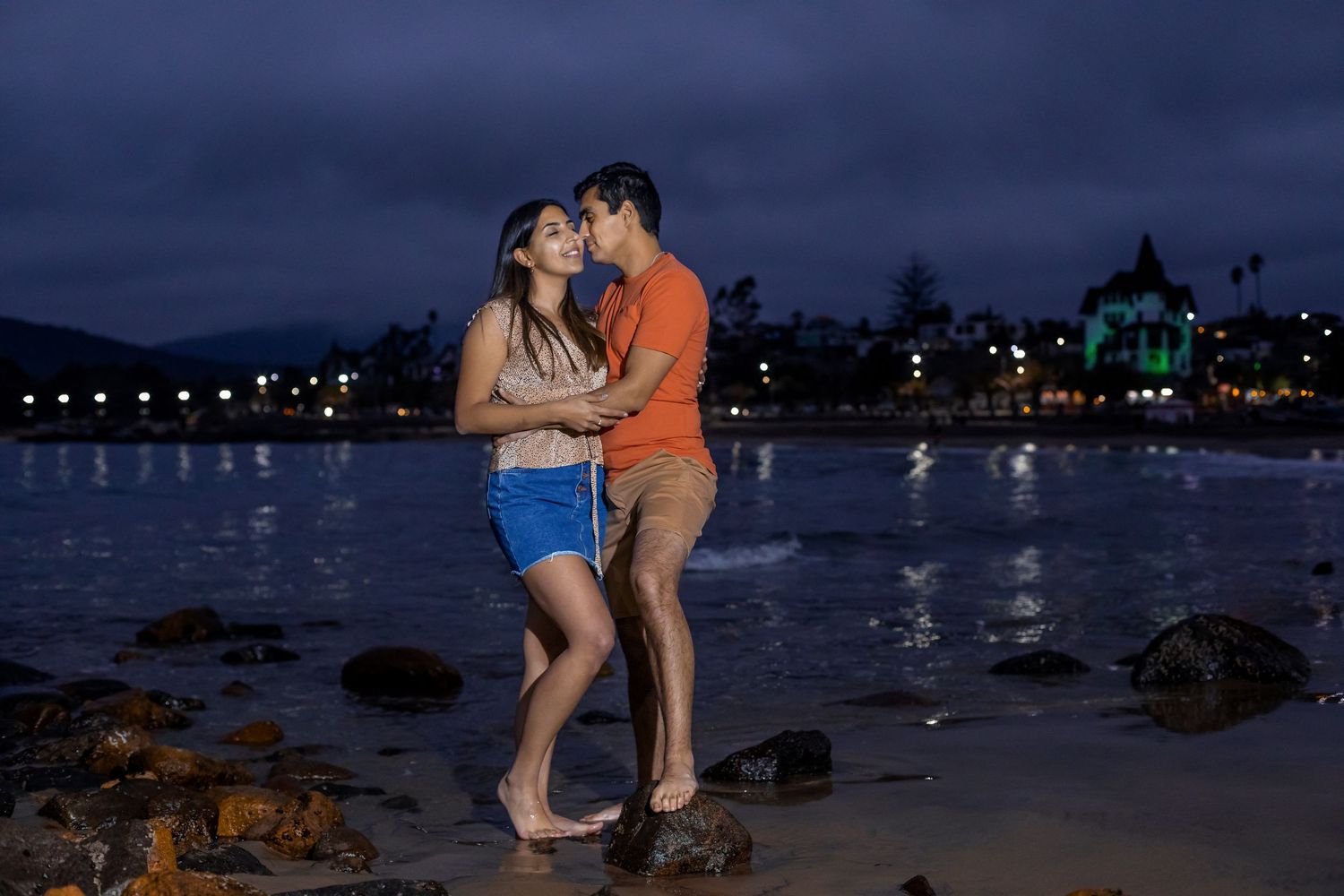 Couple sharing a romantic moment on a beach at dusk with city lights reflecting on water.