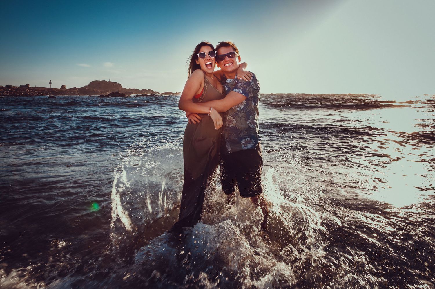 Laughing couple standing in splashing ocean waves on a sunny coastal beach.