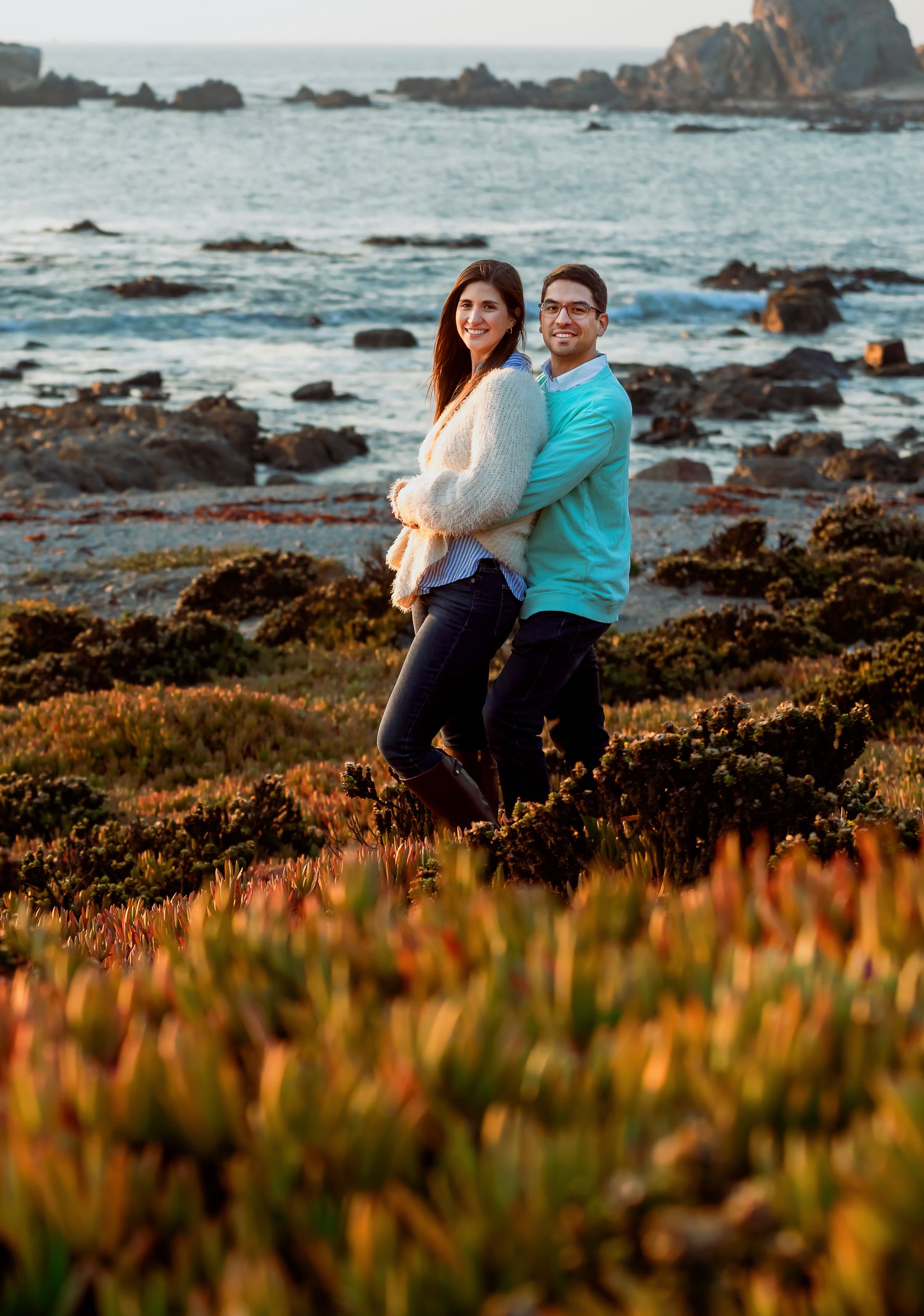 Couple embracing on rocky coastal shoreline with ocean waves and colorful succulents in golden hour light.