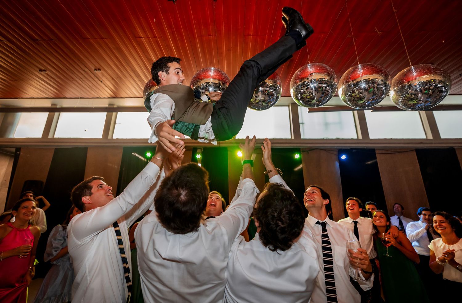 Groom being lifted and tossed in the air by groomsmen under disco balls at reception.