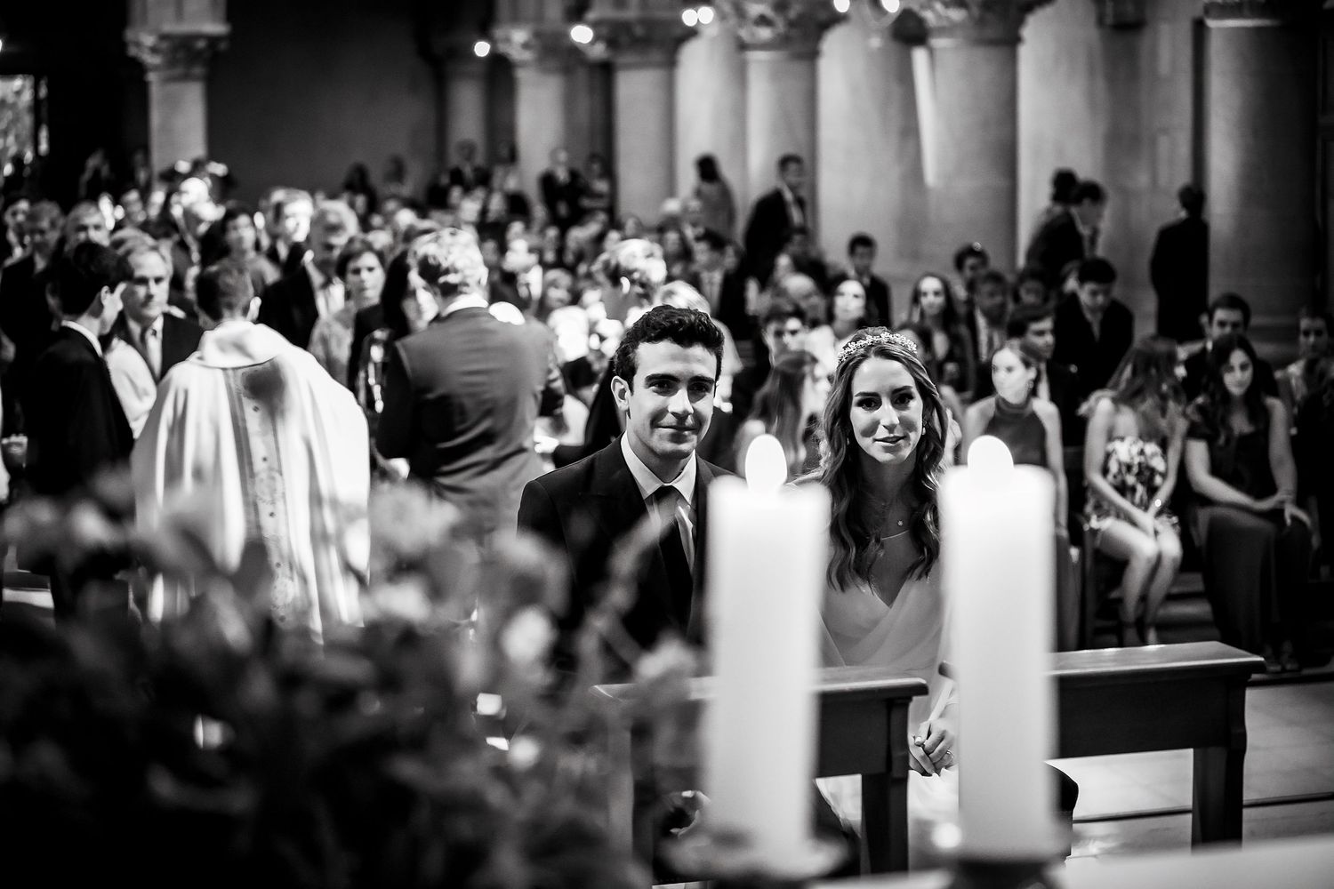 Black and white photo of bride and groom smiling at altar with candles and wedding guests in background.