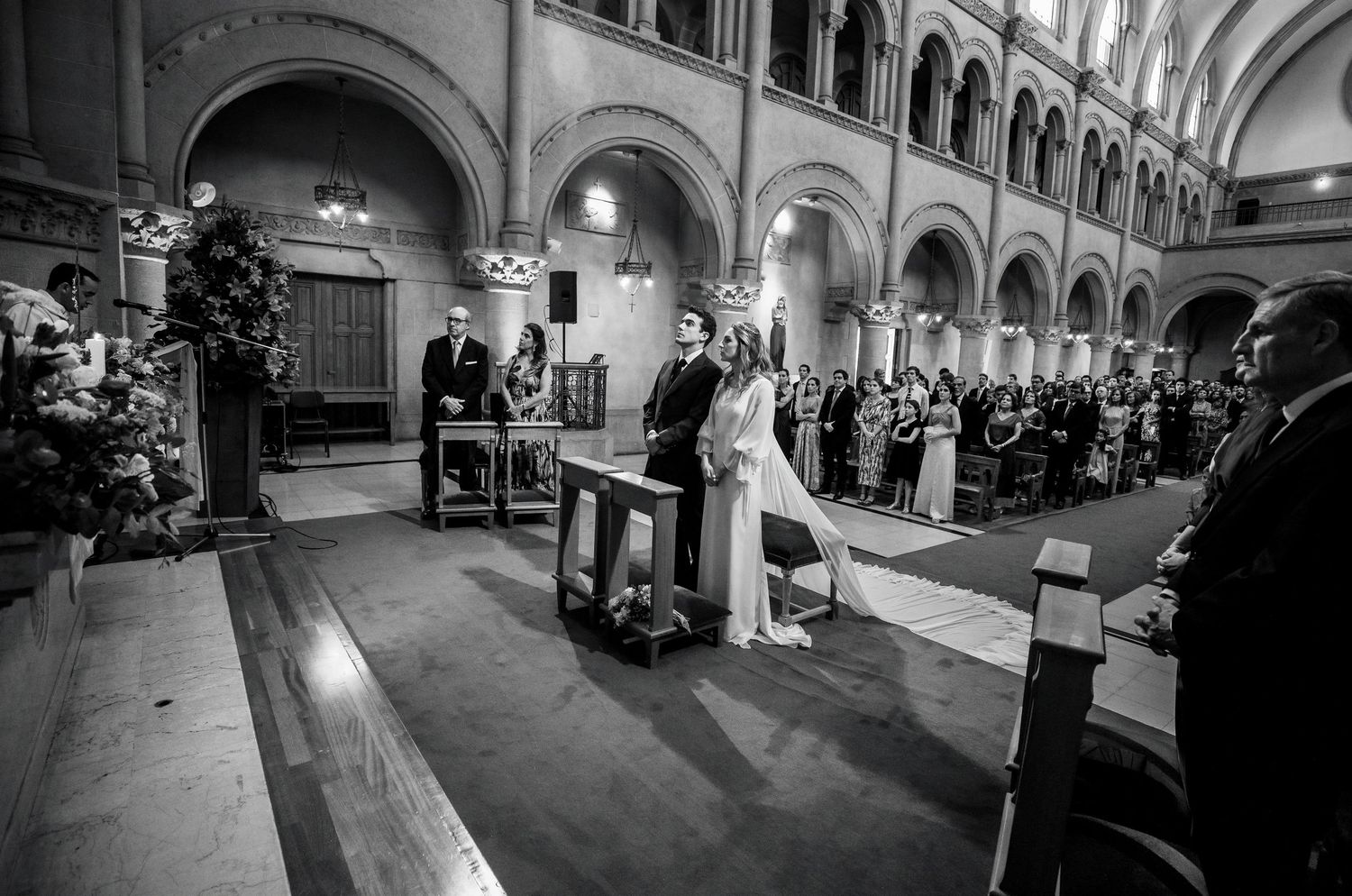 Black and white photo of wedding couple exchanging vows at church altar with guests.