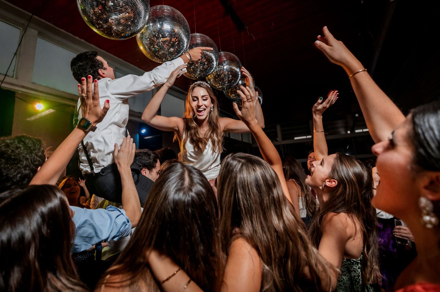 Joyful bride lifted above cheering crowd under disco balls during festive wedding reception dance celebration.