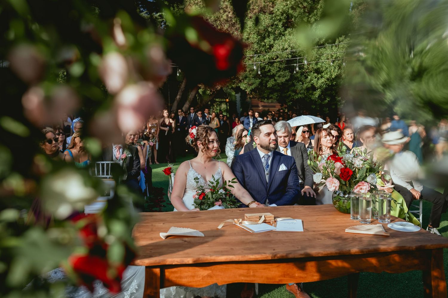 Bride and groom sit at wooden table during outdoor garden wedding ceremony framed by lush greenery.