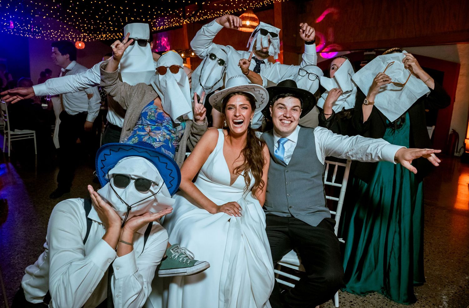 Bride and groom pose with guests wearing paper masks at fun wedding reception.