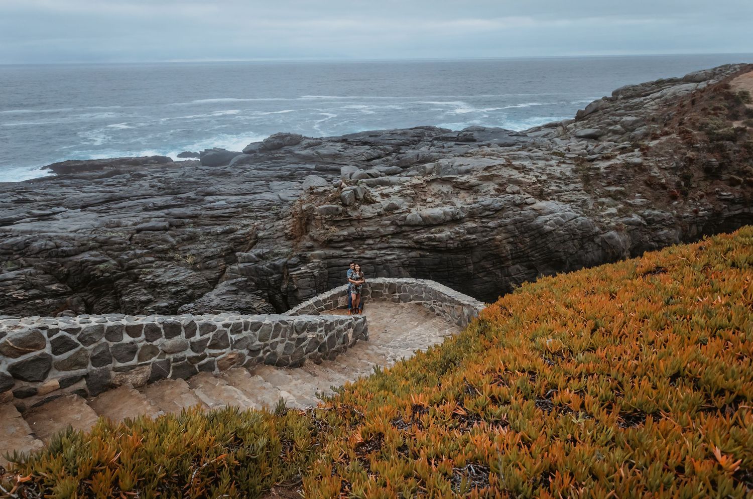 Stone wall overlook on rugged coastline with orange succulents and moody ocean views.