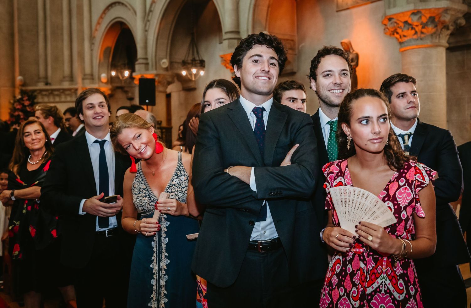 Wedding guests watching ceremony in decorated church, man in suit with arms crossed in foreground.