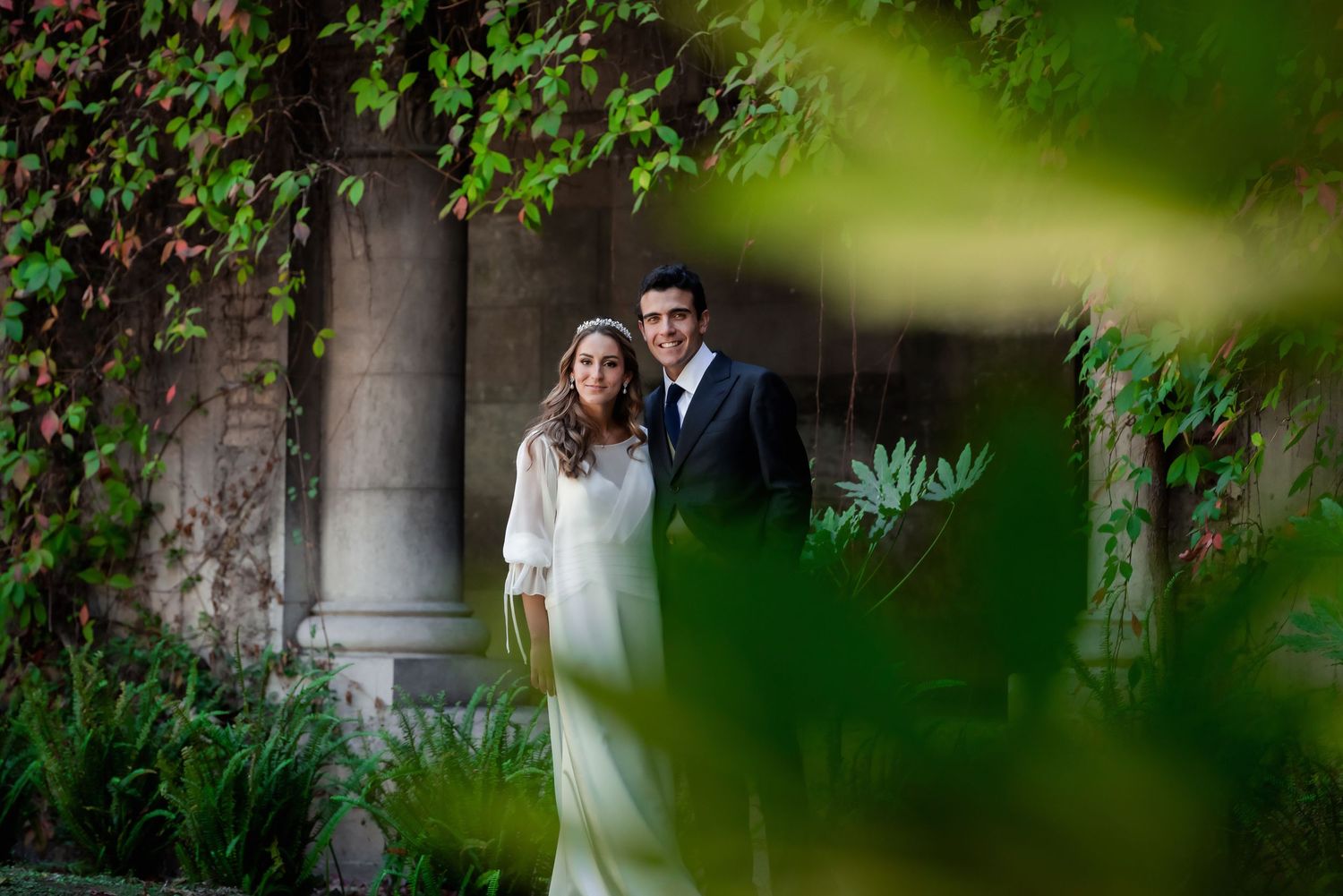 Bride and groom framed by lush green leaves posing in a garden with stone columns behind them.