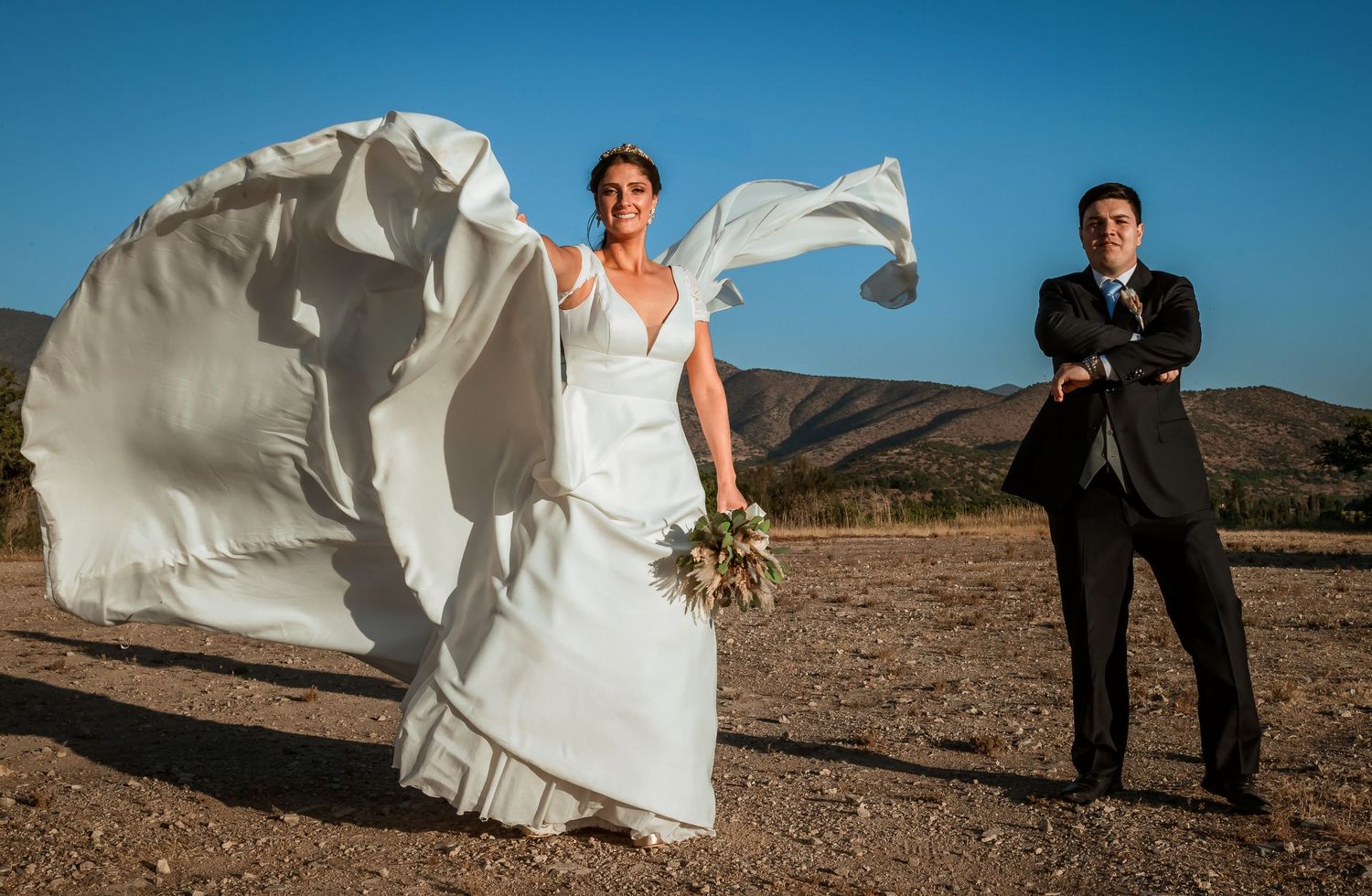 Bride's wedding dress billows dramatically in wind while groom stands confidently with arms crossed outdoors.