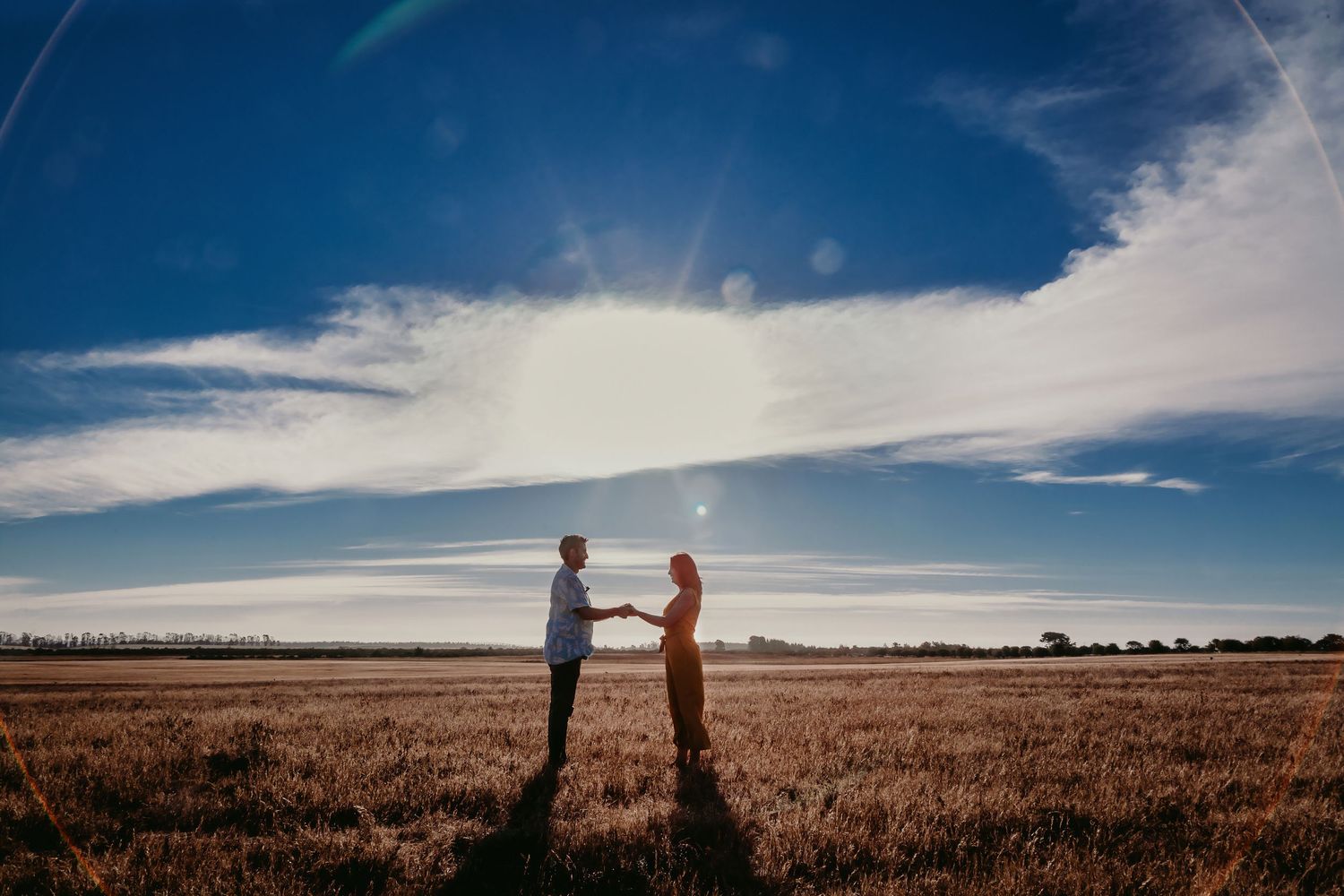Couple holding hands facing each other in a vast wheat field under a dramatic blue sky with sun rays.