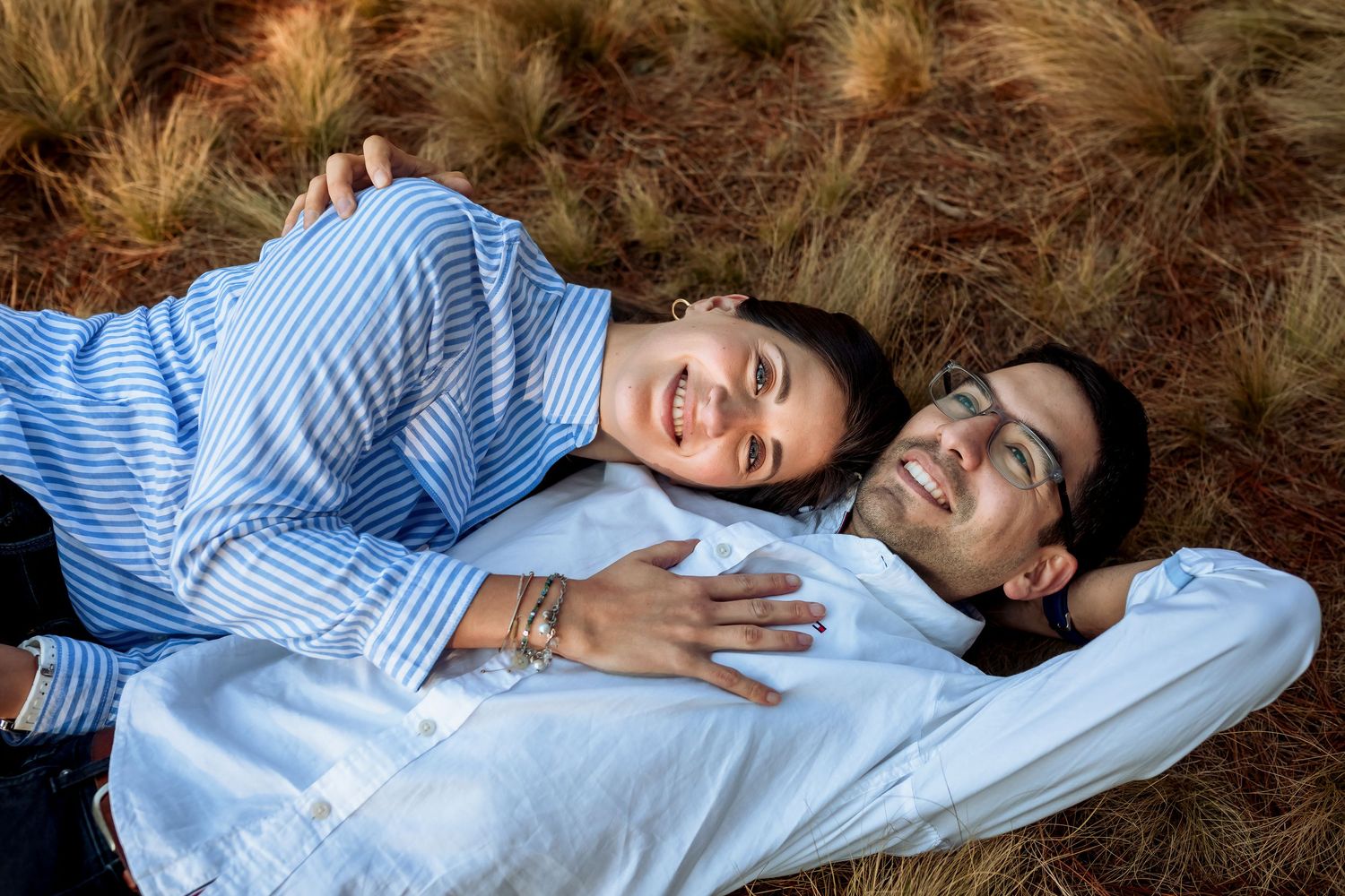 A joyful couple lying on dry grass, smiling and holding each other, viewed from above.
