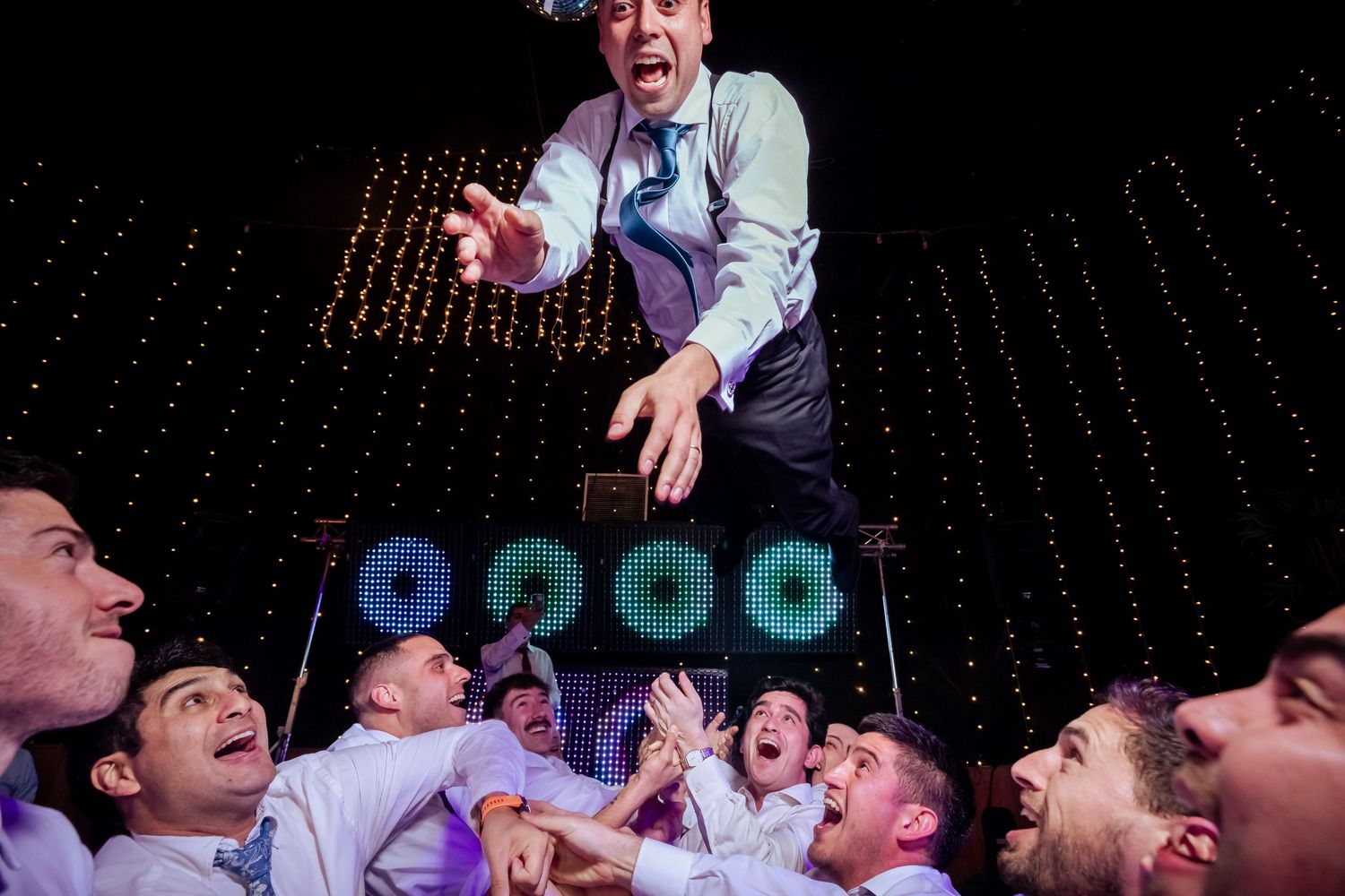 Groom is lifted by cheering groomsmen under twinkling lights at a high-energy wedding reception.