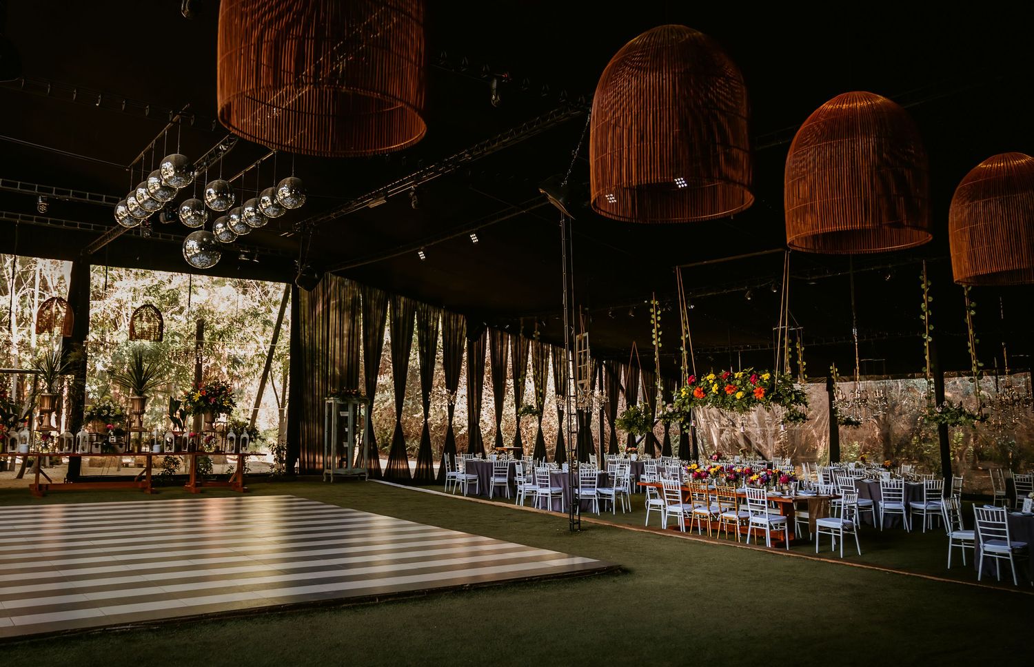 Elegant wedding reception venue with wicker pendant lights, striped dance floor, and floral centerpieces on long white tables.