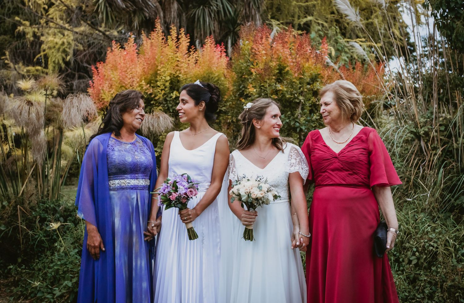 Two brides pose with their mothers holding bouquets in a colorful autumn garden setting.