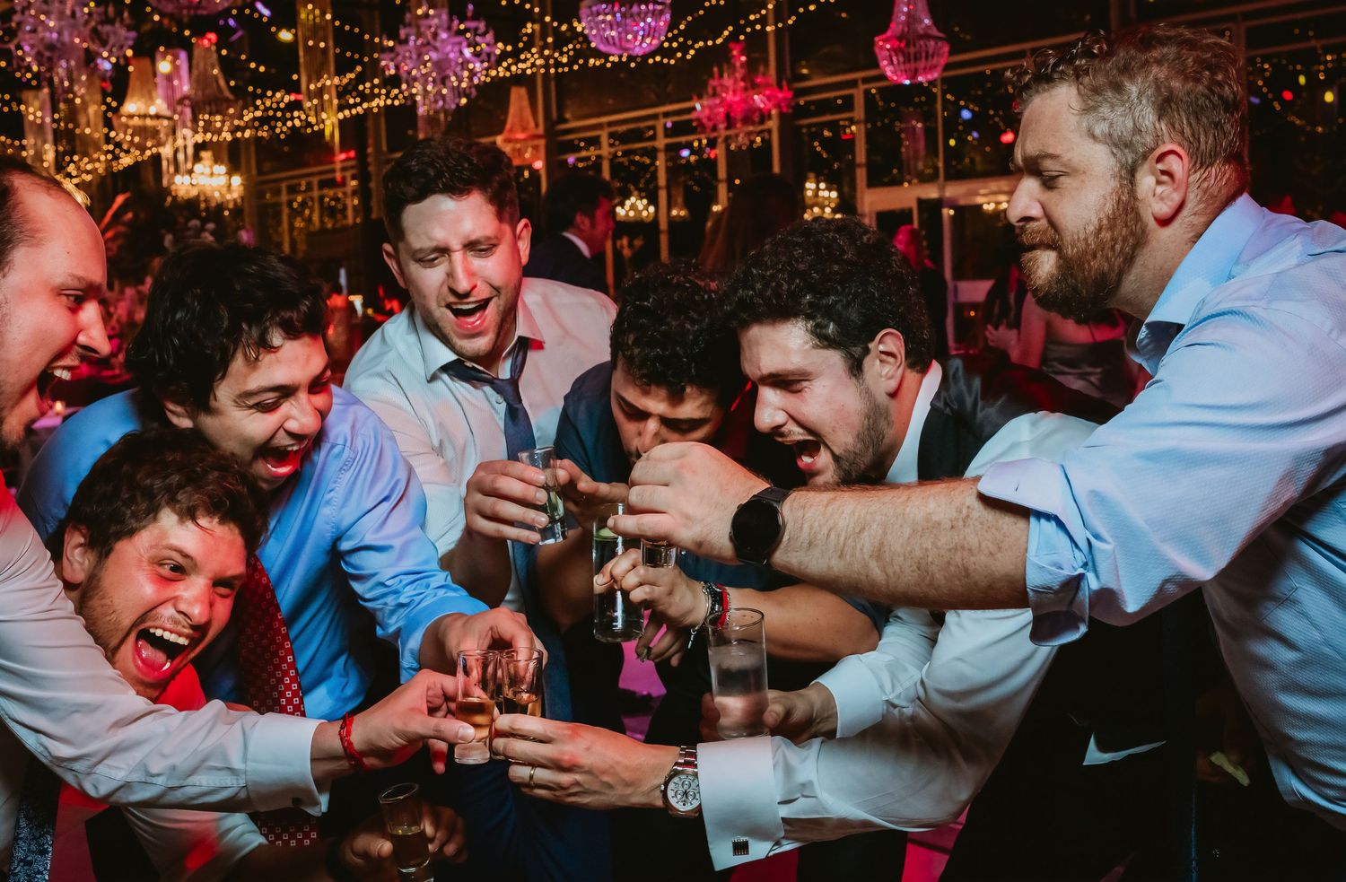 Group of men in dress shirts toasting drinks together at a lively wedding celebration.