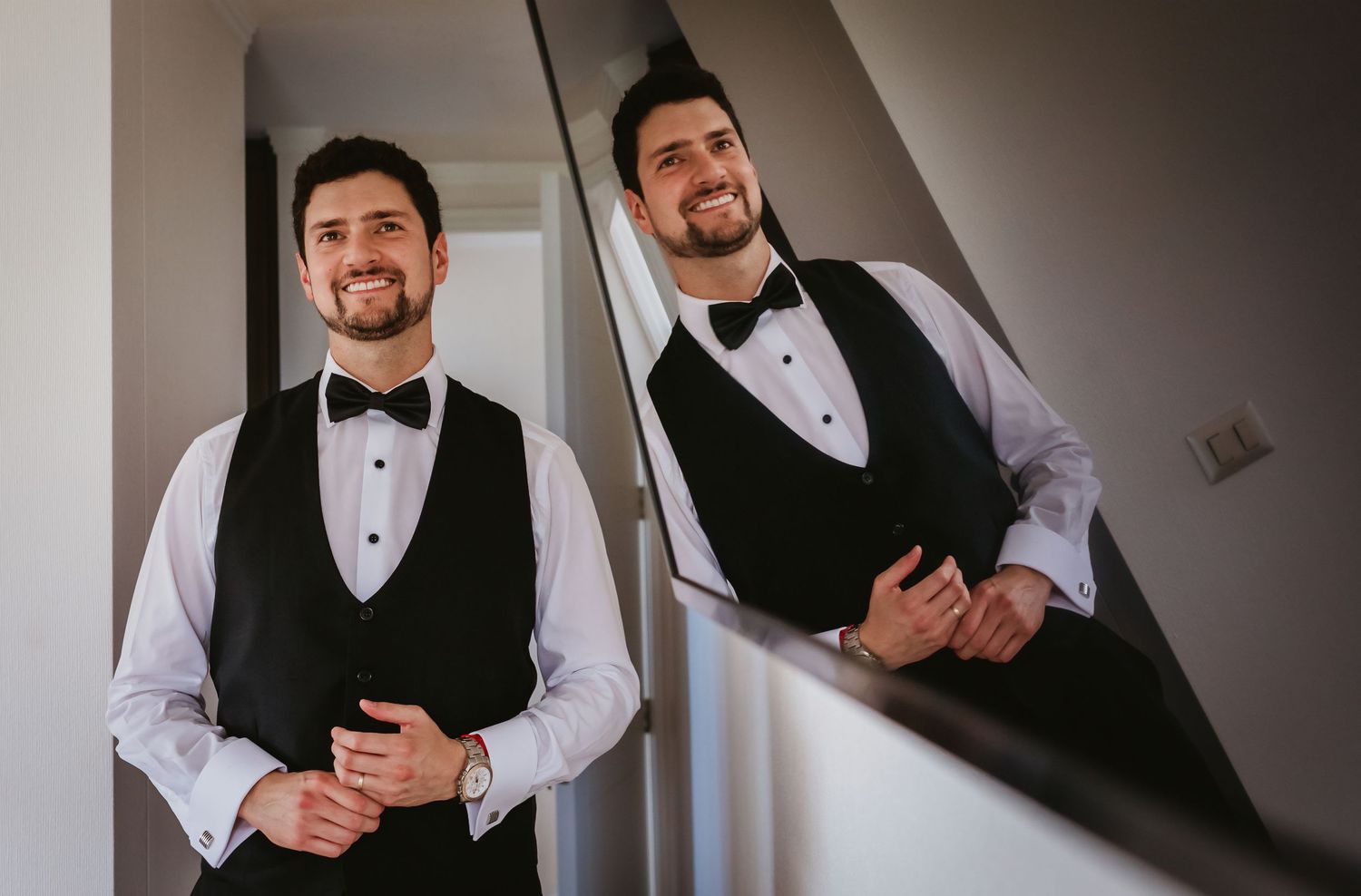 Handsome groom in black tuxedo vest and bow tie smiling at his reflection in a mirror.