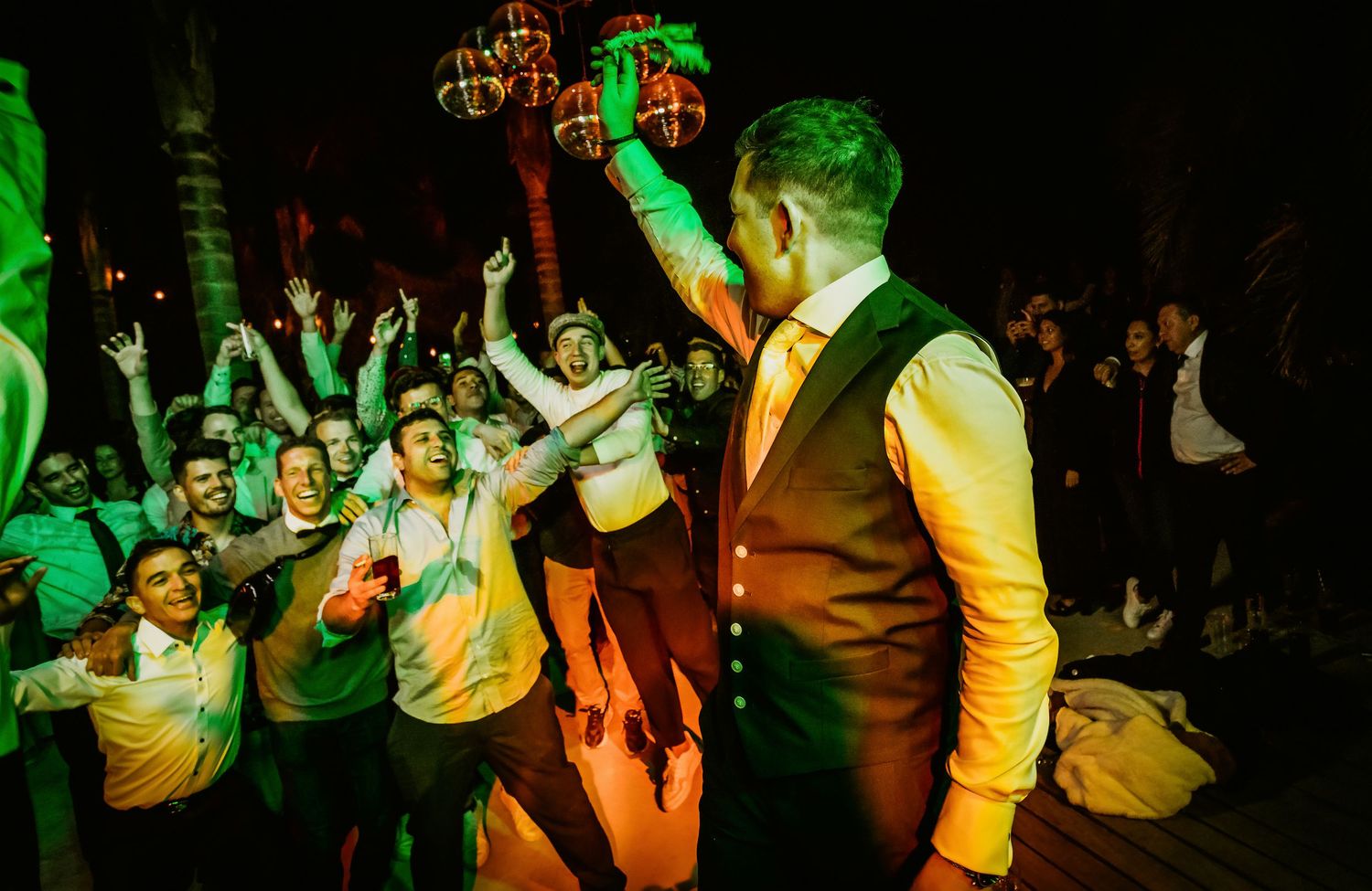 Groom raising bottle to cheering crowd of wedding guests on lively outdoor dance floor at night.