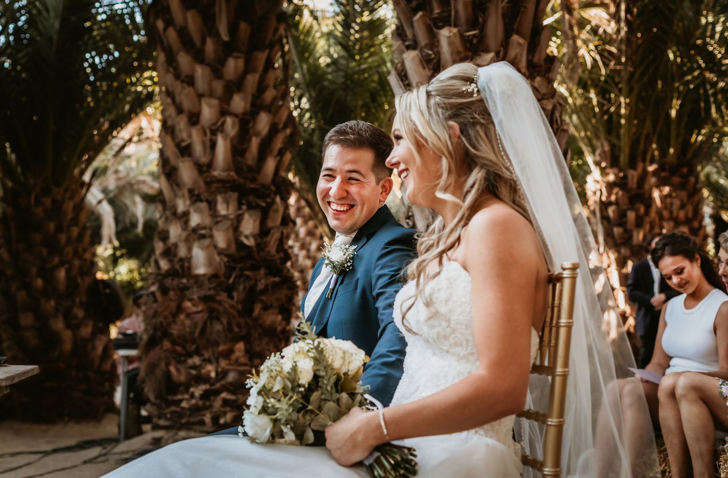 Laughing bride holding white bouquet and groom in navy suit share joyful moment at wedding ceremony.