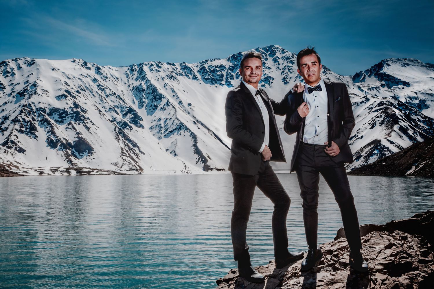 Two grooms in black tuxedos pose playfully beside a stunning alpine lake with snowy peaks.