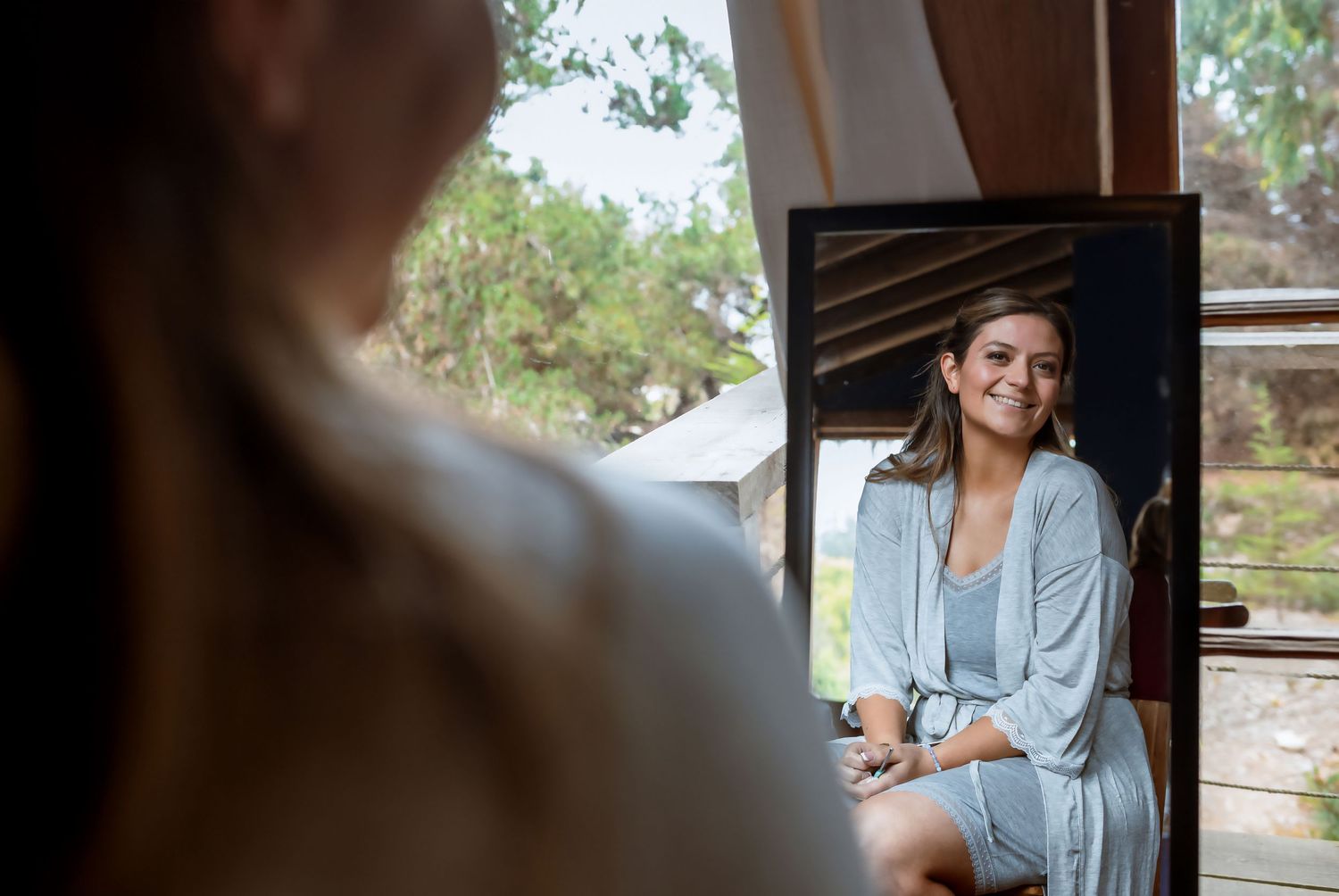 Smiling bride in blue robe looking at her reflection in mirror while getting ready.