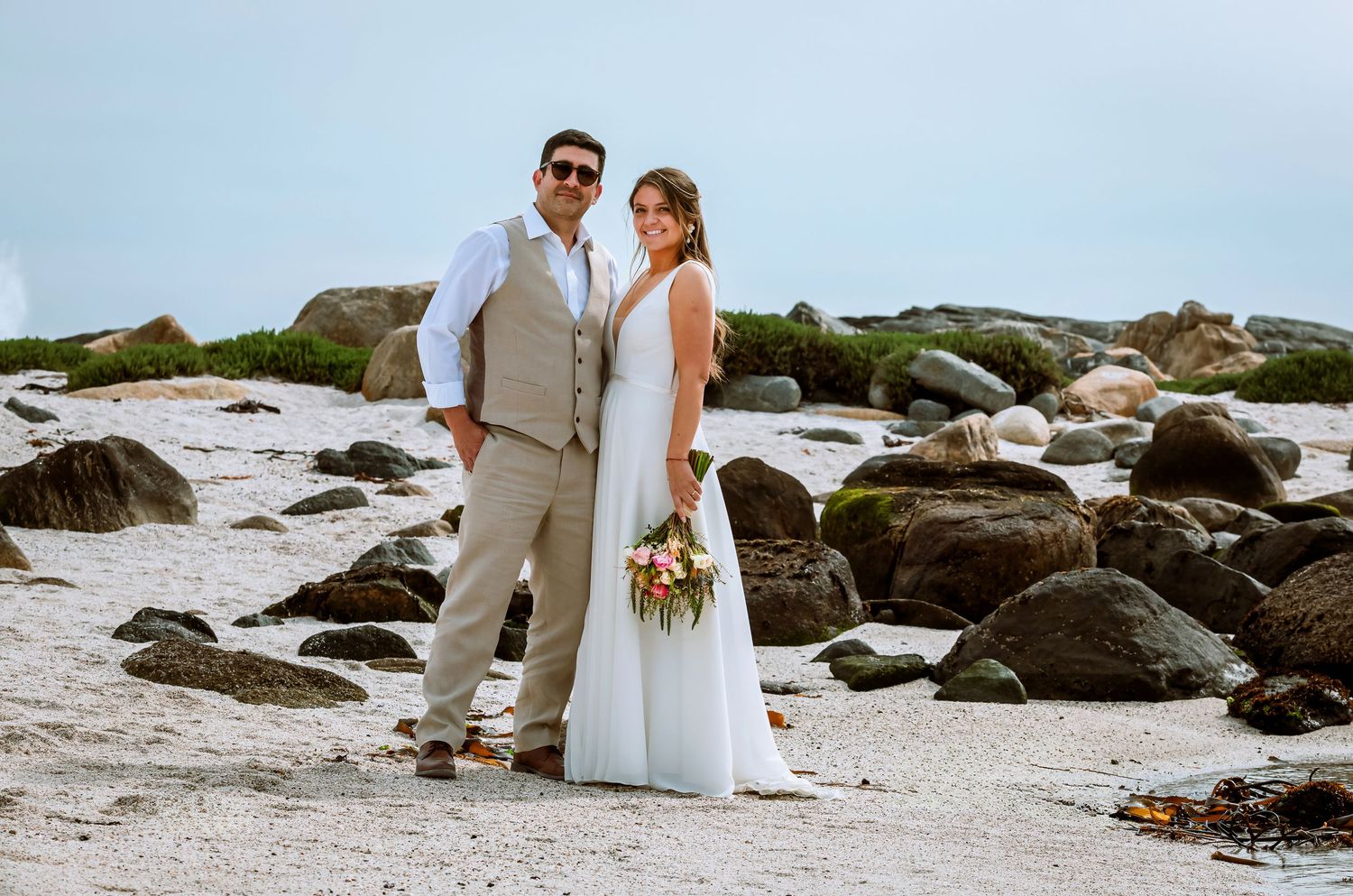 Bride holding bouquet and groom in tan suit posing together on sandy beach with large rocks behind them.