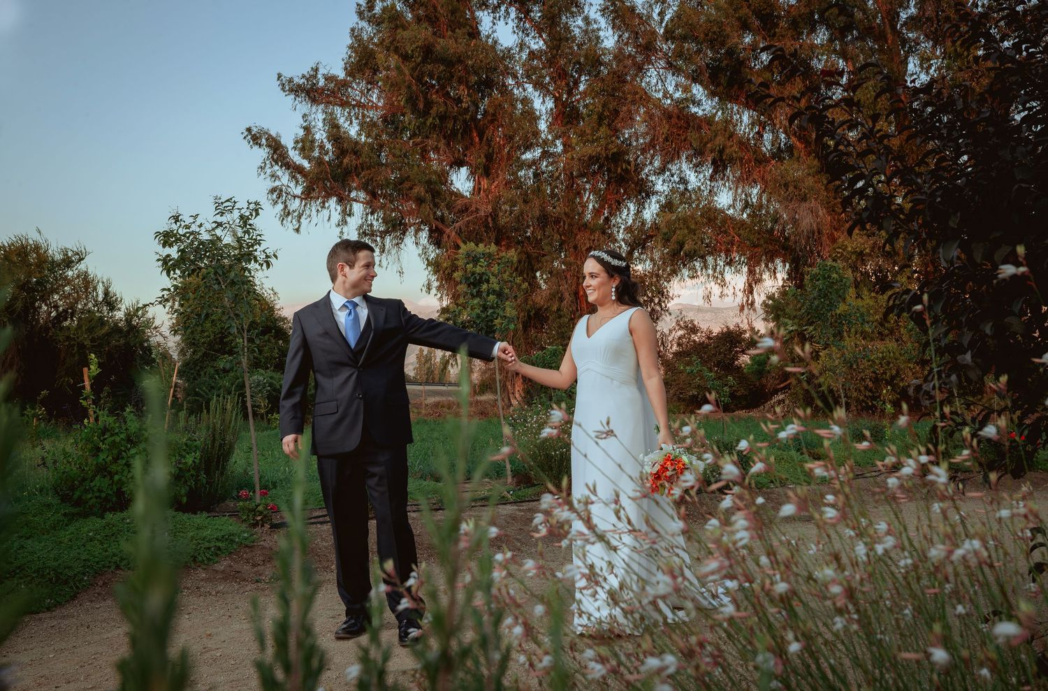 Wedding couple strolling hand in hand through a colorful garden with warm sunset lighting.