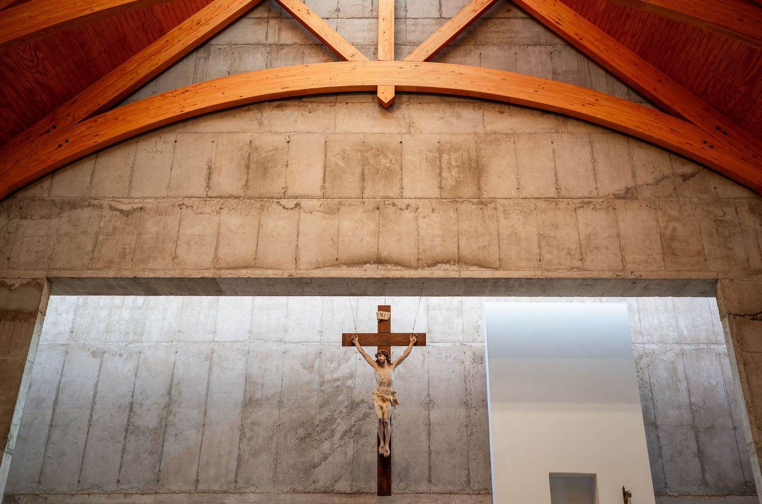 Wooden arched ceiling church interior with crucifix on concrete wall above altar.