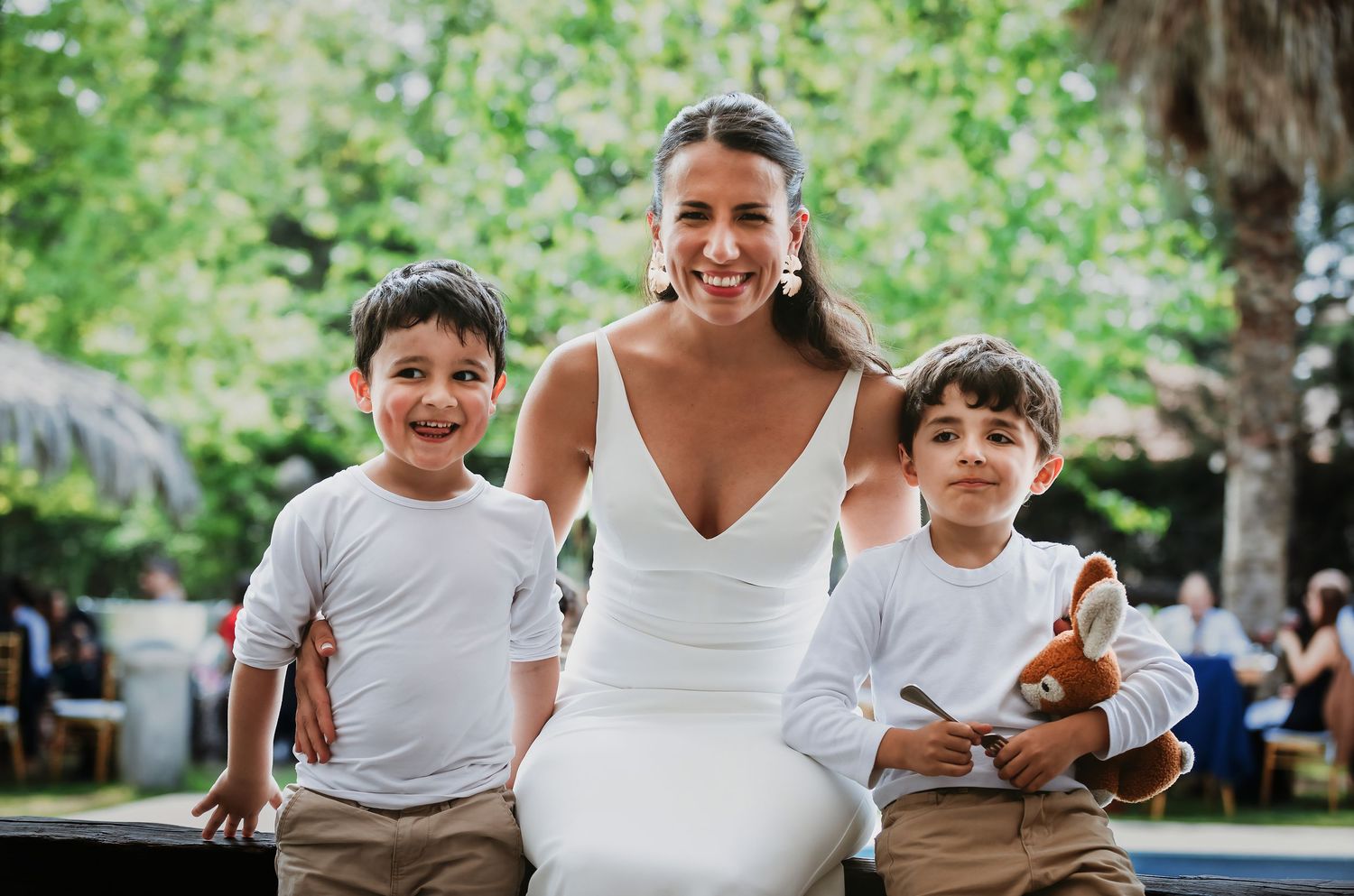 Smiling bride in white dress poses with two young boys in white shirts and khaki pants outdoors.