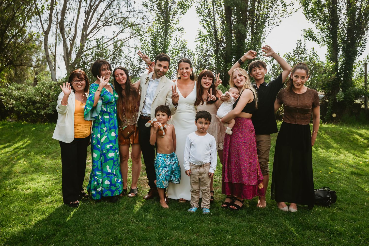 Wedding party group photo on green lawn with bride, groom, family and friends posing joyfully outdoors.