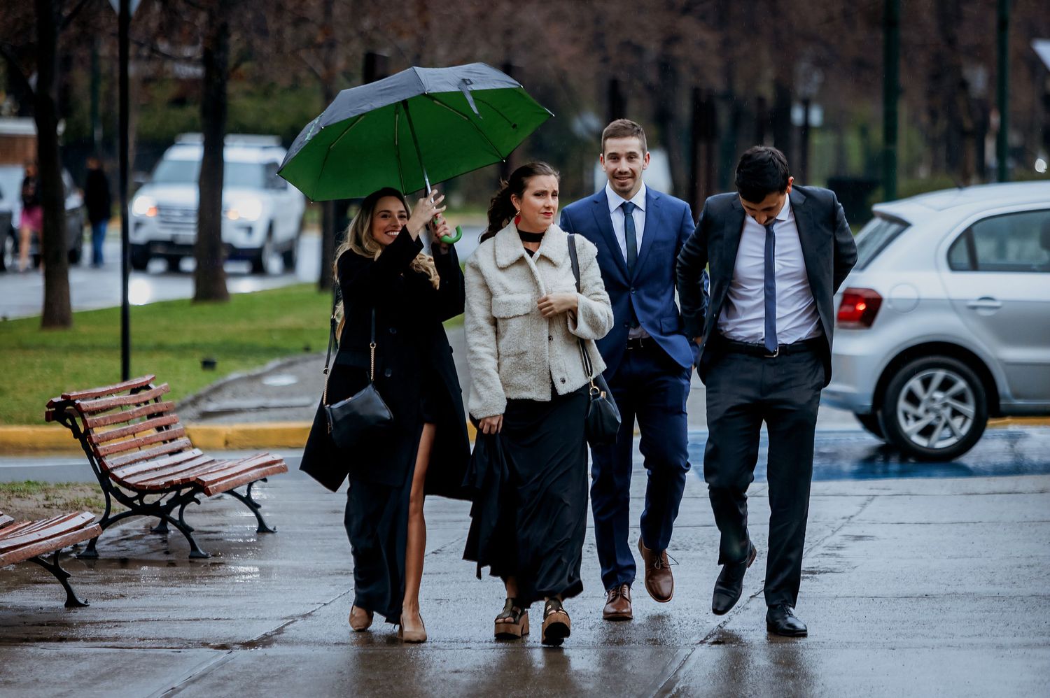 Four well-dressed people walking outdoors in the rain, one holding a green umbrella on a wet city path.