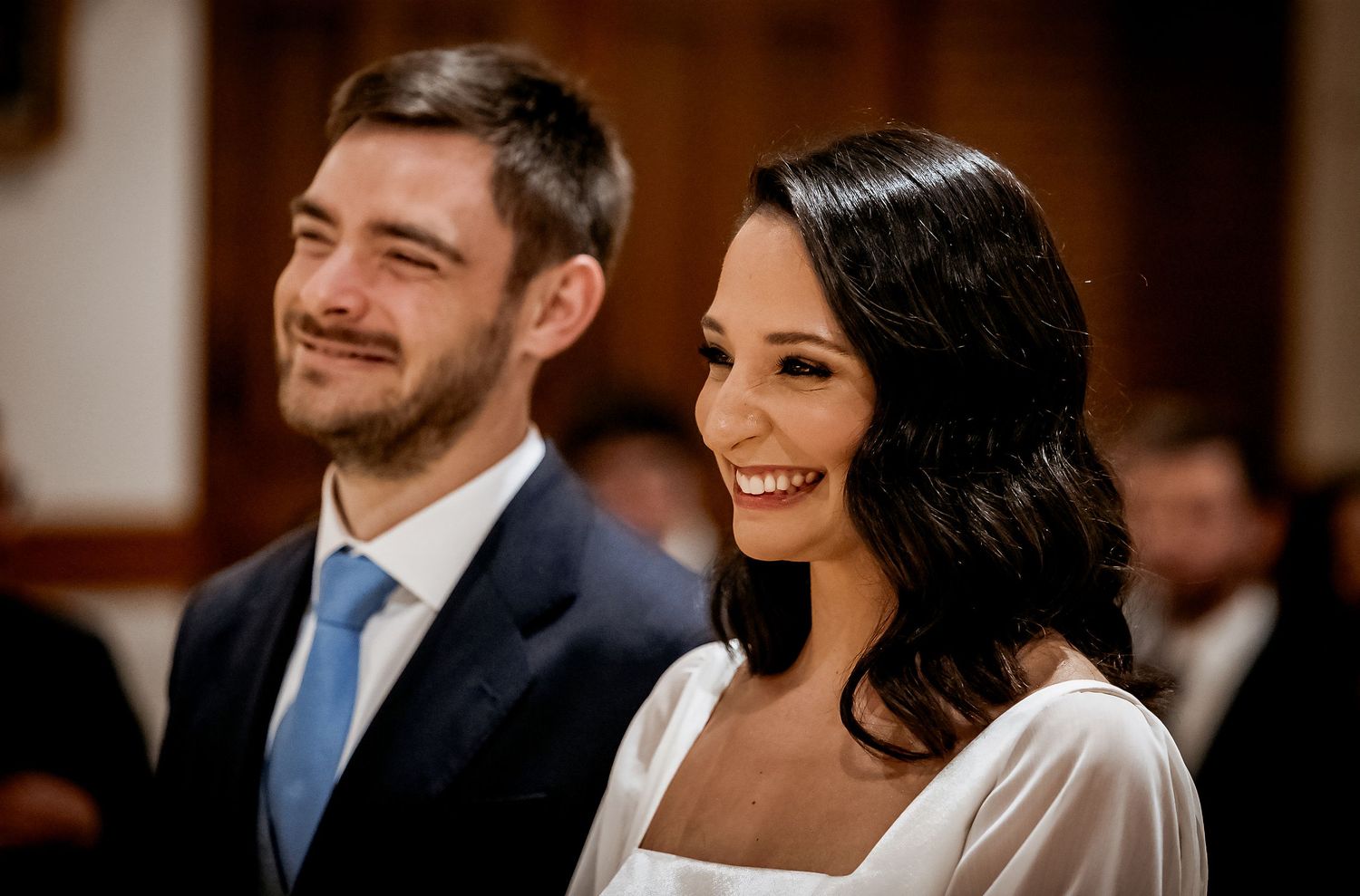 Happy couple smiling at their wedding ceremony, bride in white dress and groom in navy suit with blue tie.