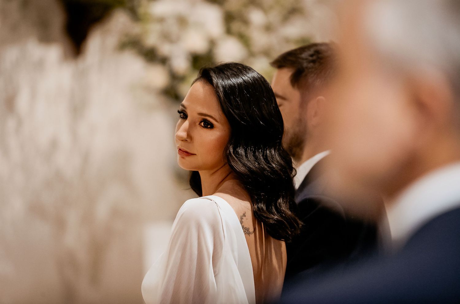 Bride in white gown glancing back during wedding ceremony with guests in soft bokeh background.