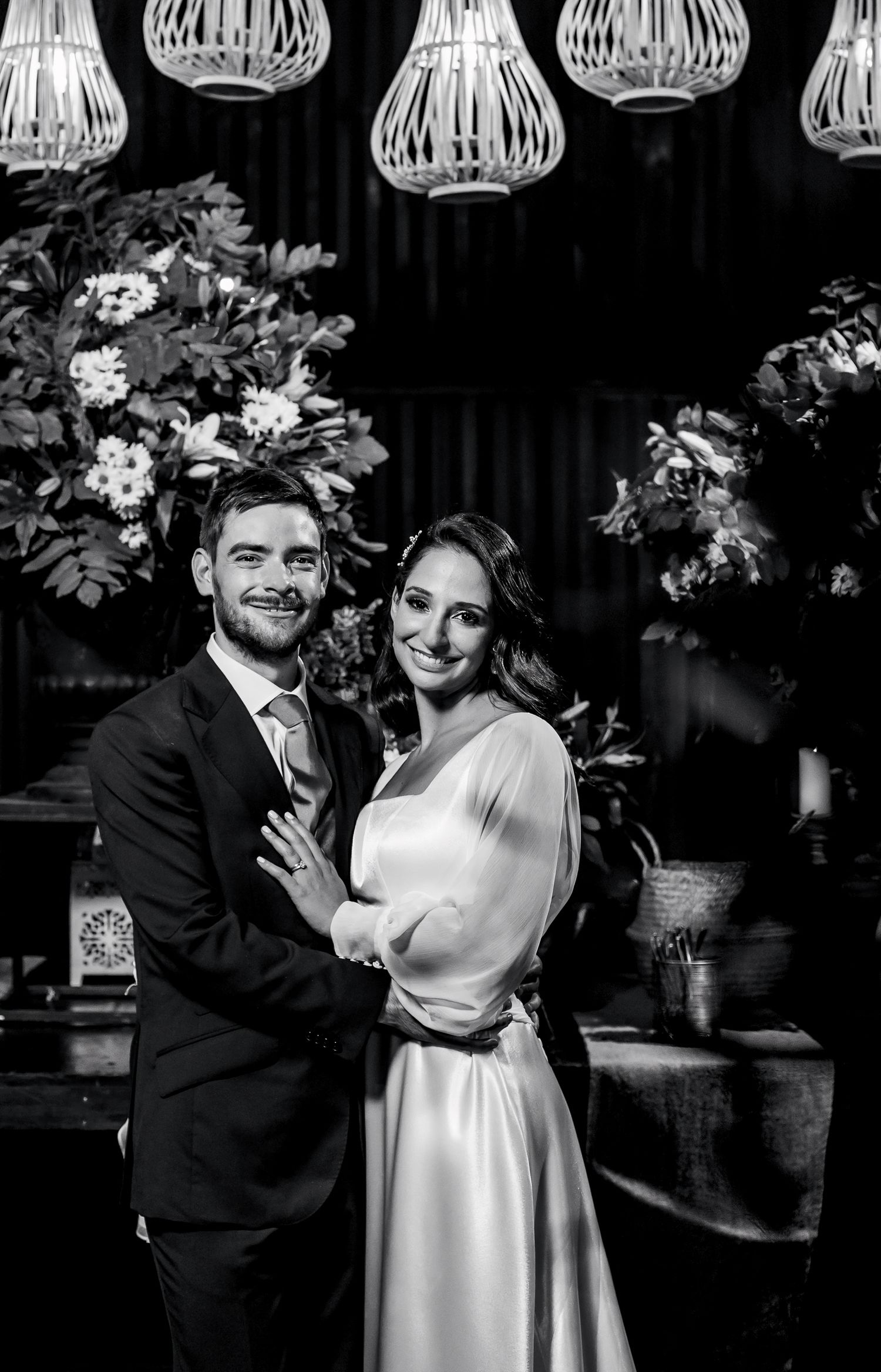 Black and white wedding photo of smiling couple embracing, bride in white gown, groom in suit, floral backdrop.