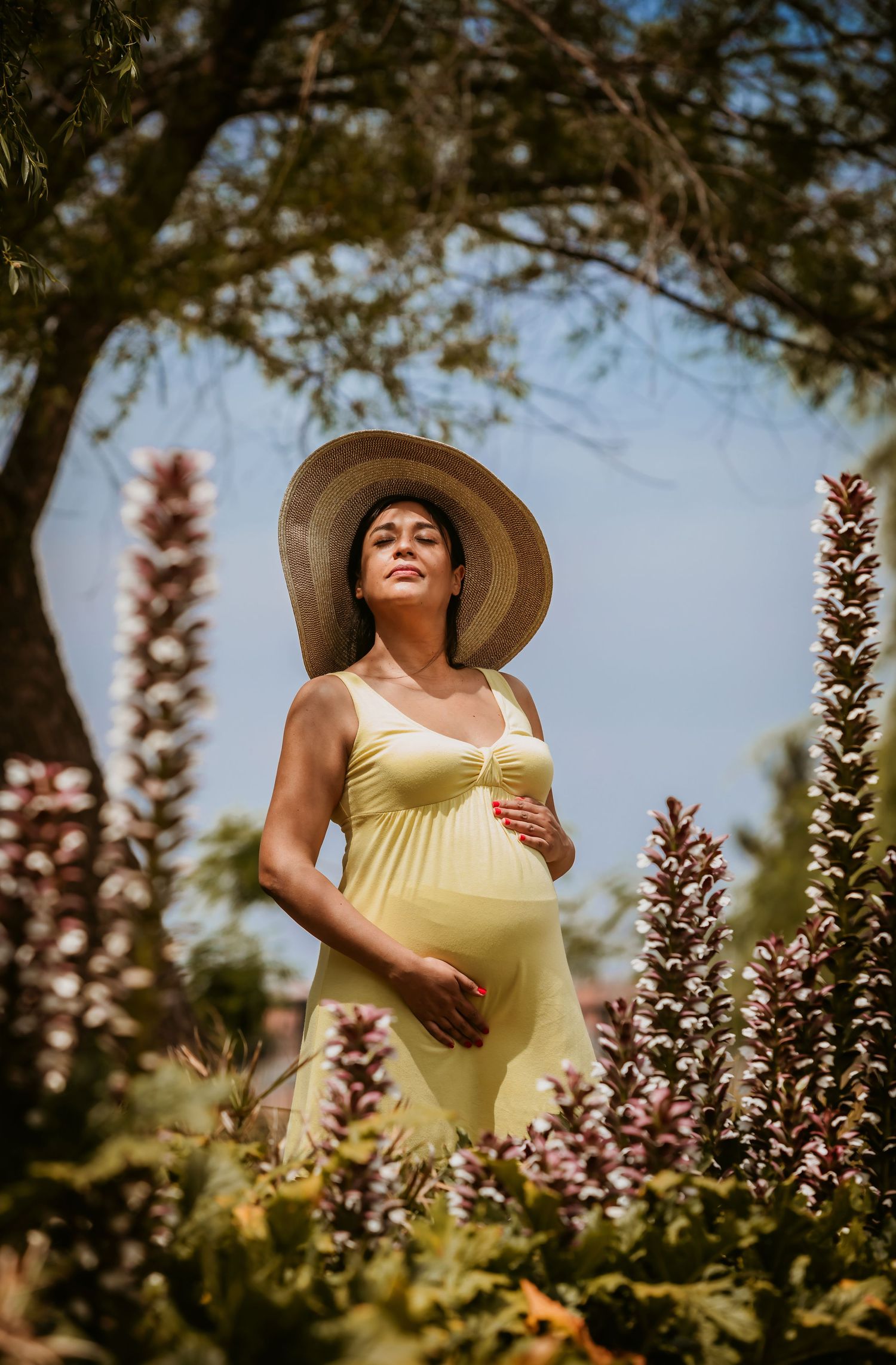 Pregnant woman in yellow dress and straw hat stands among purple flowers in a sunny garden.