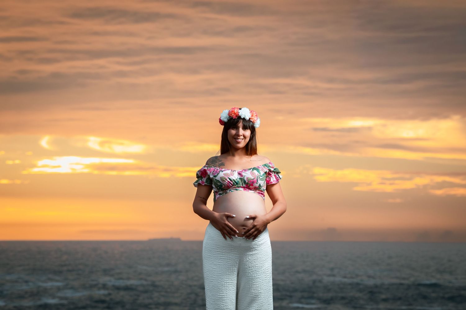 Happy pregnant woman in floral off-shoulder top and flower crown poses at ocean sunset.