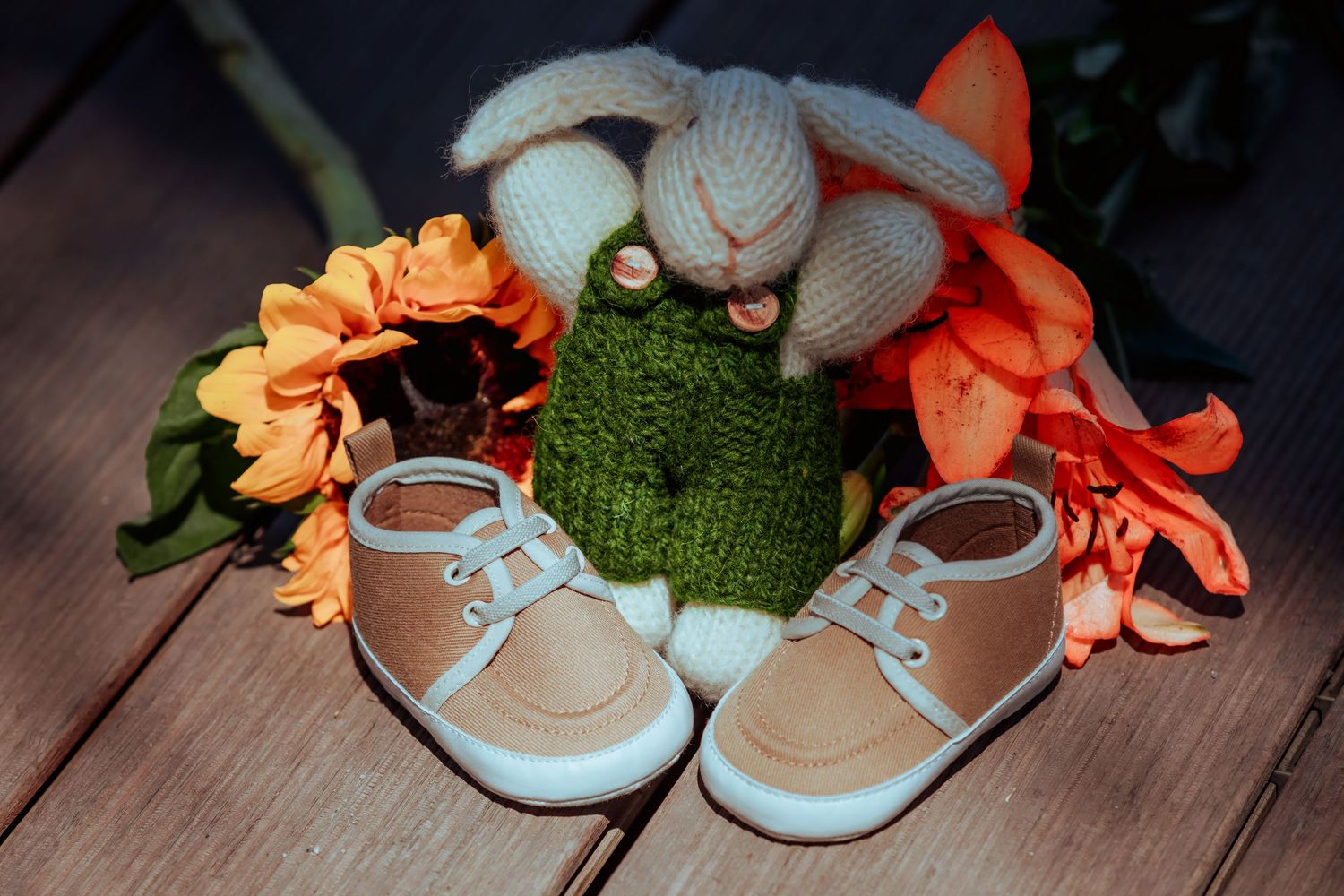 Tiny baby shoes beside knitted bunny and frog toys with orange flowers on a wooden surface.