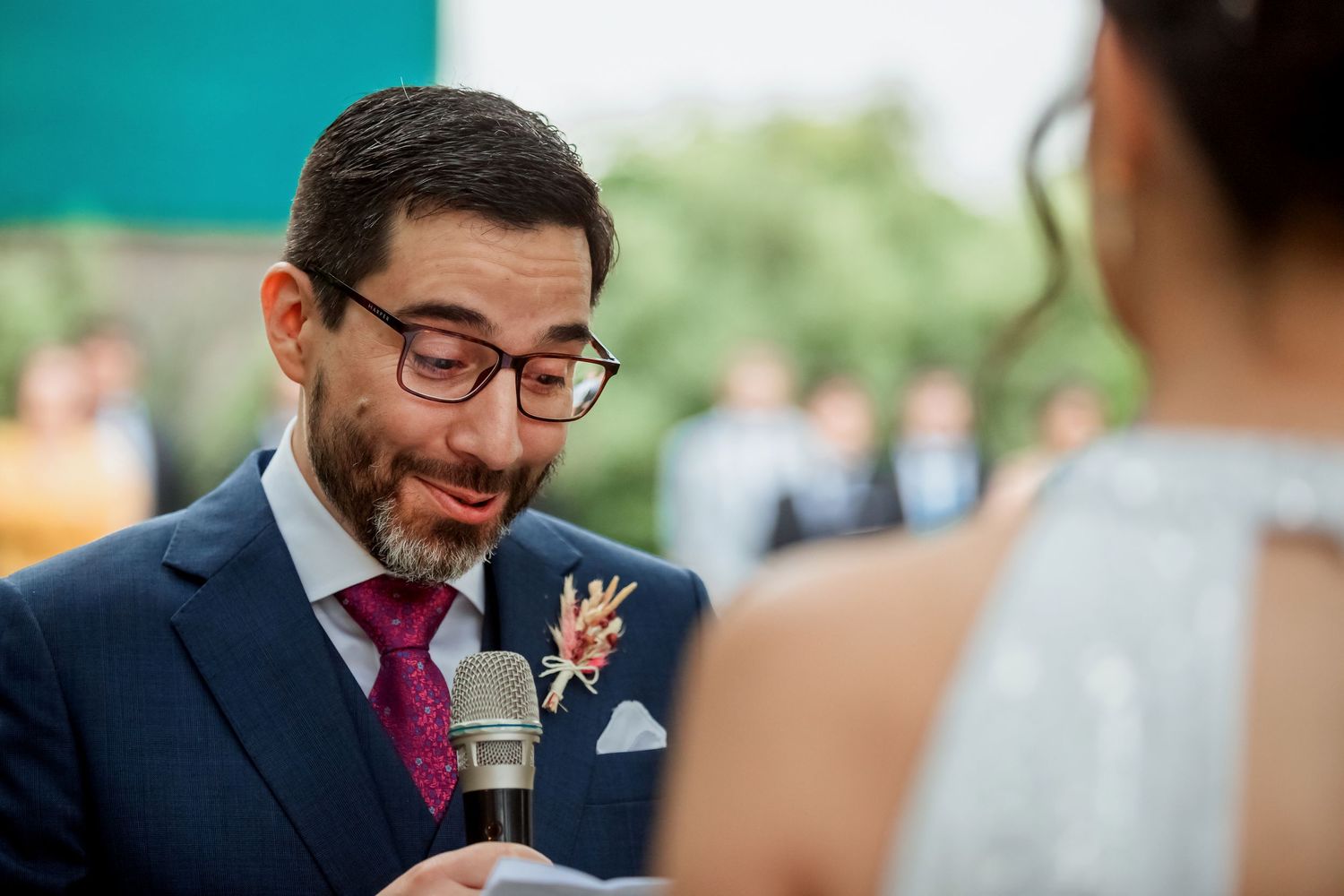 Emotional groom in navy suit with burgundy tie listens to wedding vows outdoors.