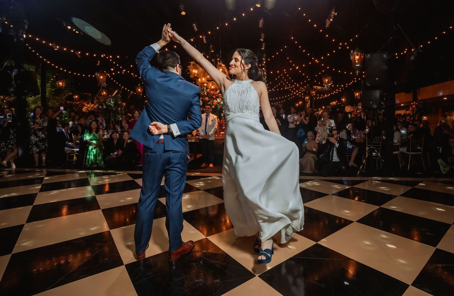 Groom spins bride in white gown on checkered dance floor under warm string lights.