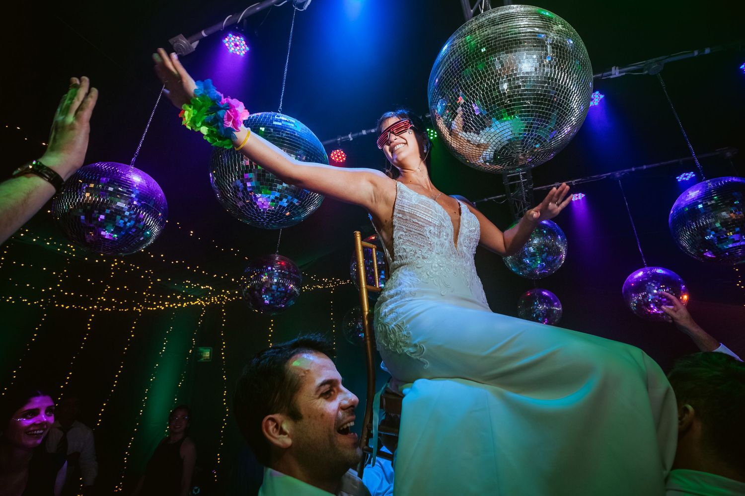 Bride lifted by groom on dance floor surrounded by disco balls at vibrant wedding reception party.