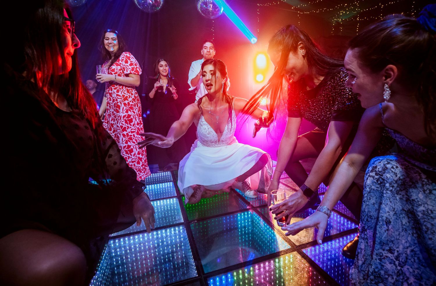 Bride laughing joyfully on illuminated dance floor surrounded by bridesmaids at vibrant wedding reception party.