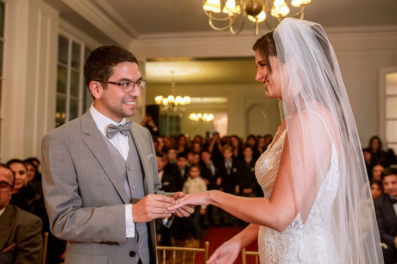 Bride placing wedding ring on groom's finger during indoor ceremony with guests seated in background.