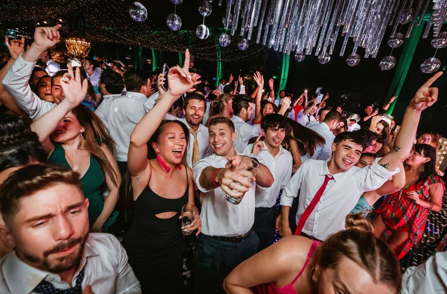 Large crowd of wedding guests dancing and celebrating with hands raised at reception party.