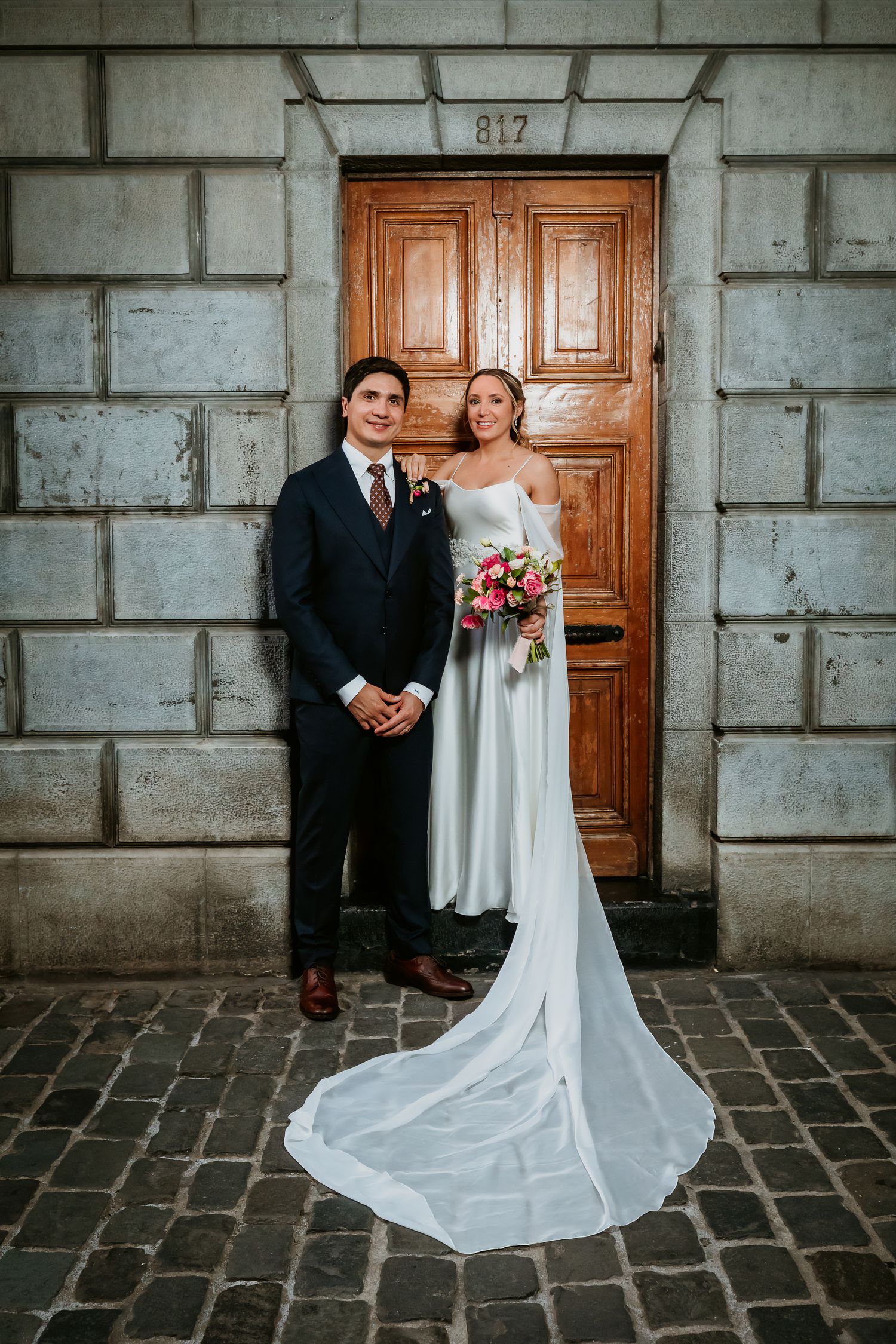 Wedding couple posing before wooden door 817, bride in white gown with long train holding bouquet.